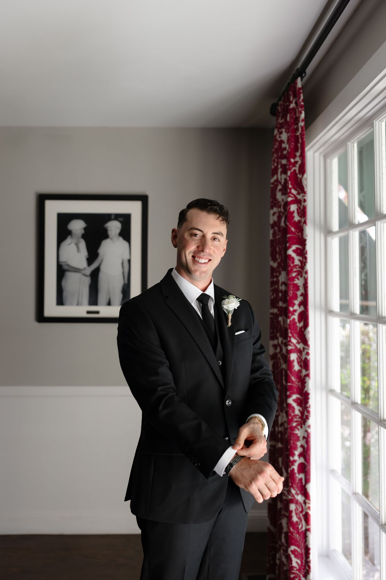 A person in a classic black suit poses near windows with red floral curtains and black and white artwork on the wall.