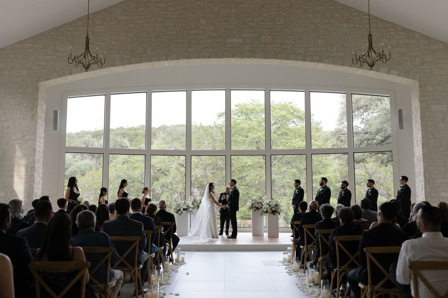 Wedding ceremony in a bright white chapel with large arched windows overlooking a scenic outdoor view.