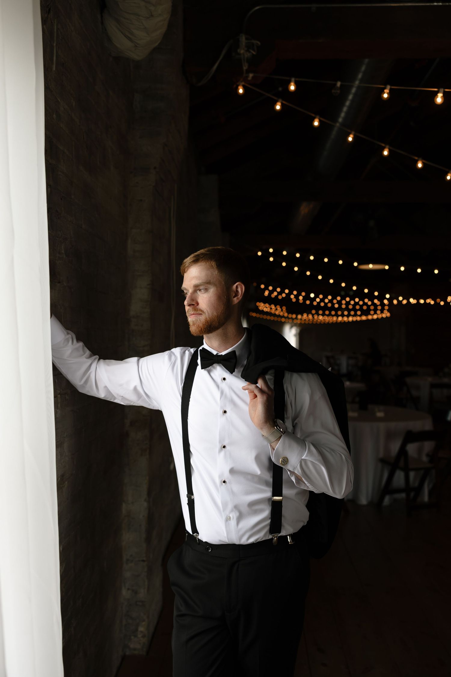 A groom in a classic white tuxedo shirt and suspenders poses in dramatic lighting against white columns and string lights.