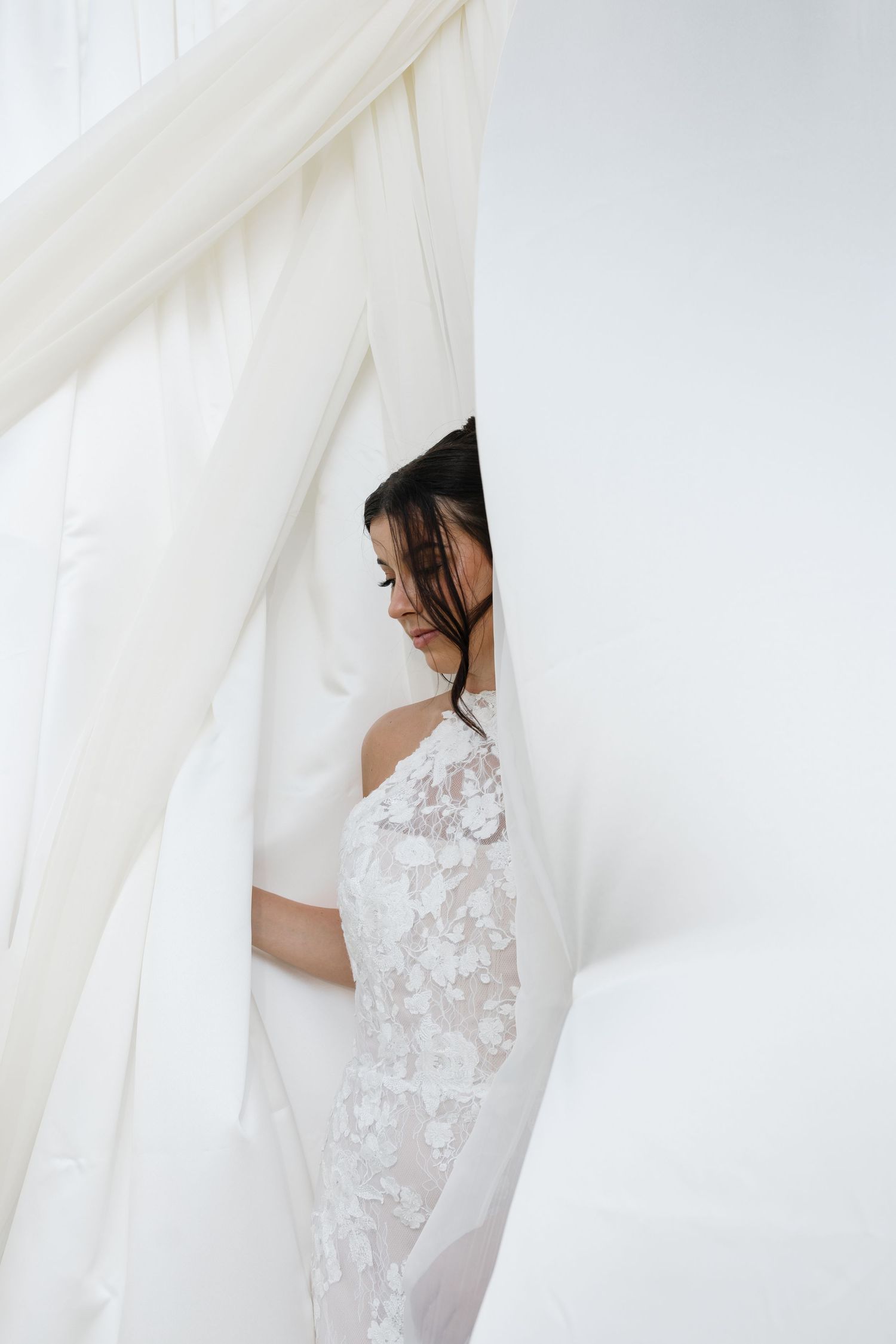 A bride in a delicate white lace wedding dress stands behind flowing sheer curtains in a bright, ethereal setting.