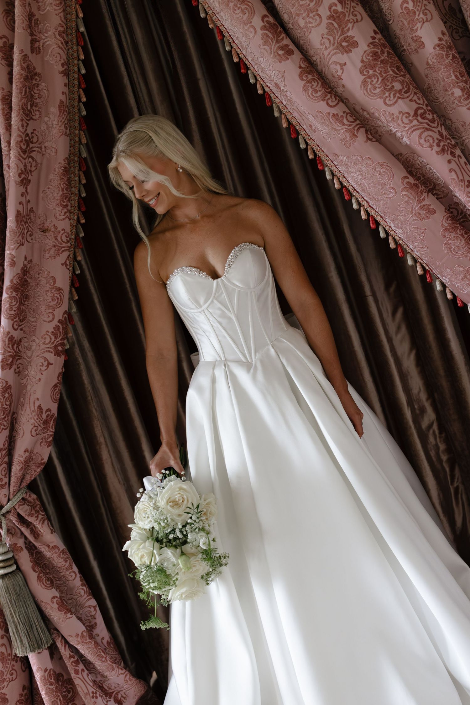 Close-up details of a white satin wedding dress with delicate lace trim and floral bouquet against mauve curtains.