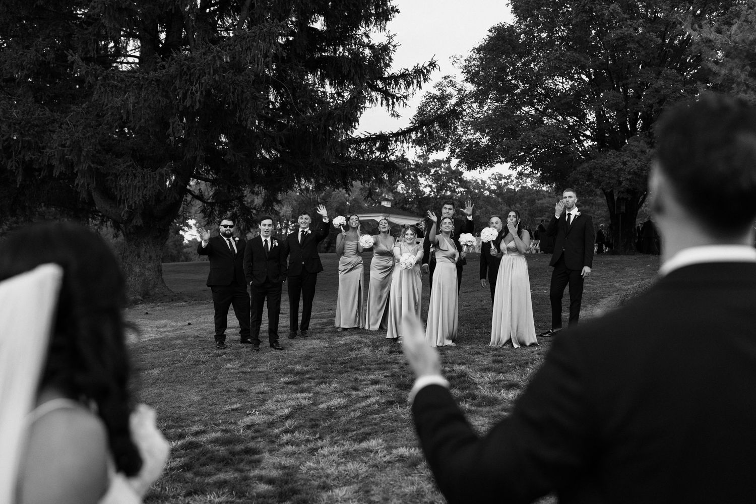 A black and white wedding party stands in a line outdoors with trees in the background.