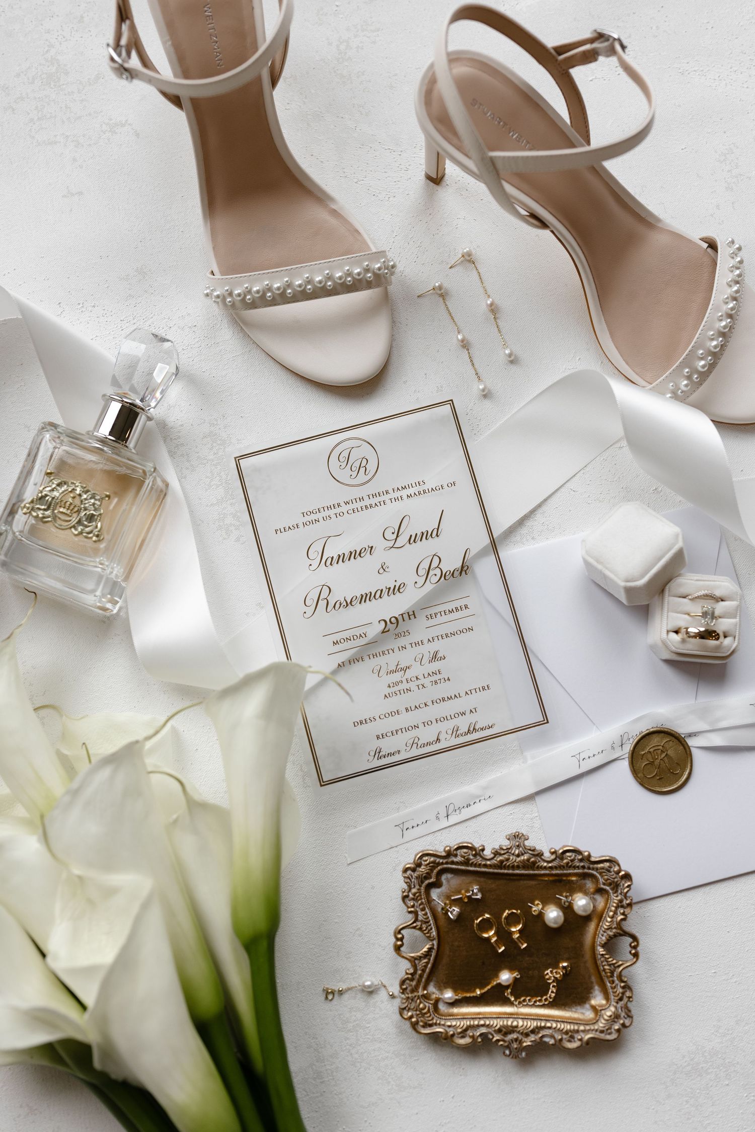 Elegant wedding details including white shoes, perfume bottles, invitations and calla lilies arranged on white surface.