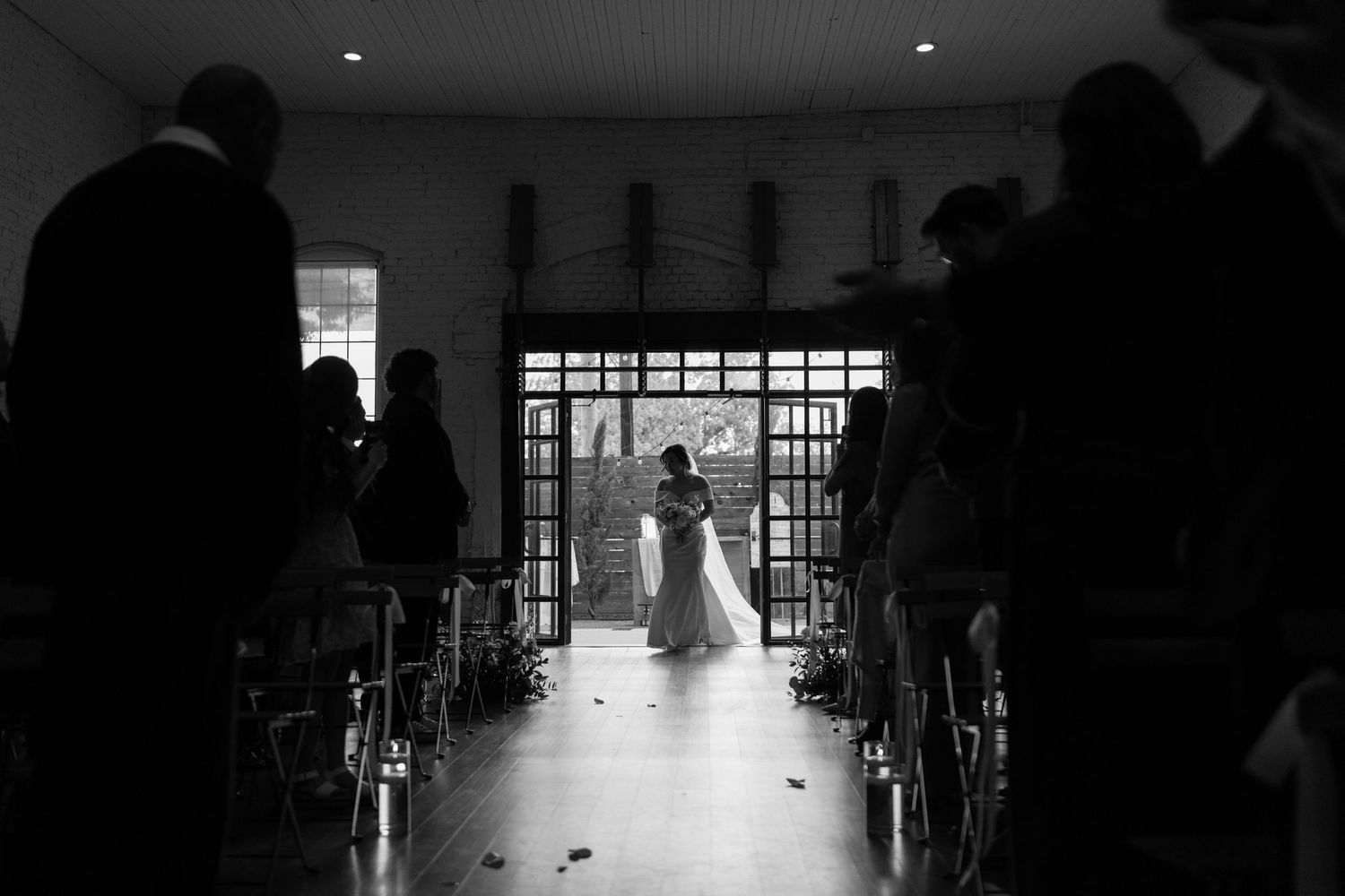 Black and white wedding photo of bride walking down the aisle toward large windows in an elegant chapel.