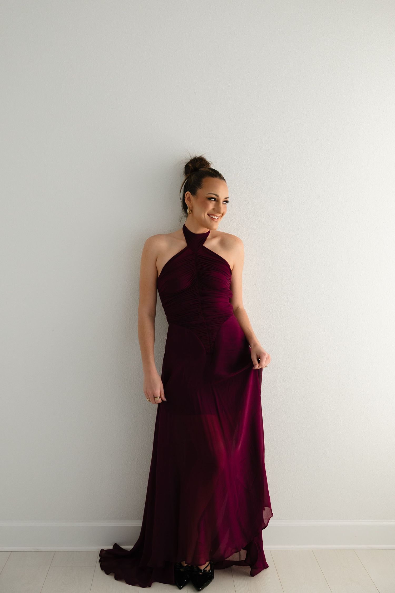 A person poses in a flowing burgundy formal dress against a white wall in a series of elegant shots.