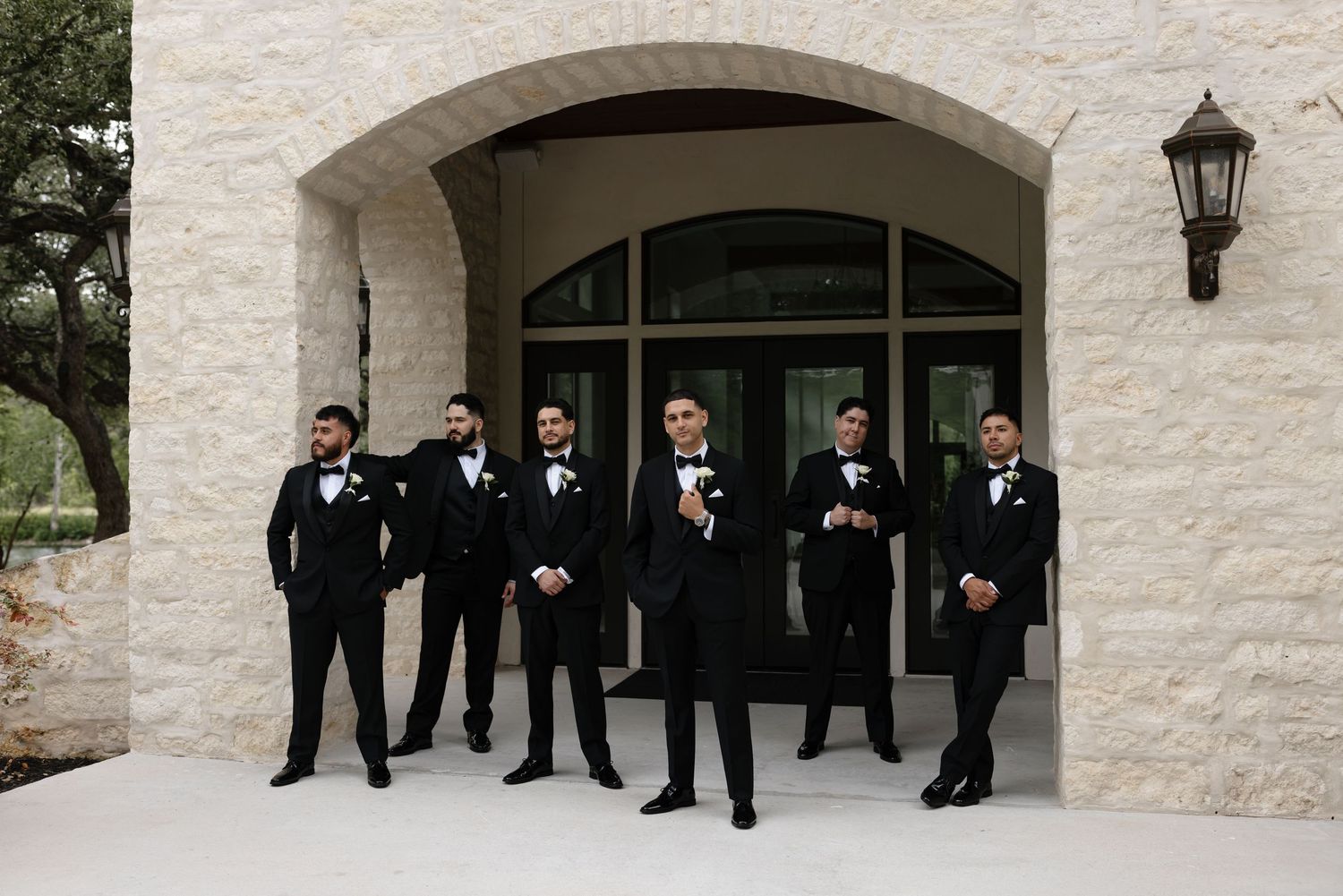 A group of groomsmen in black tuxedos stand together under a stone archway entrance with a lantern mounted on the wall.
