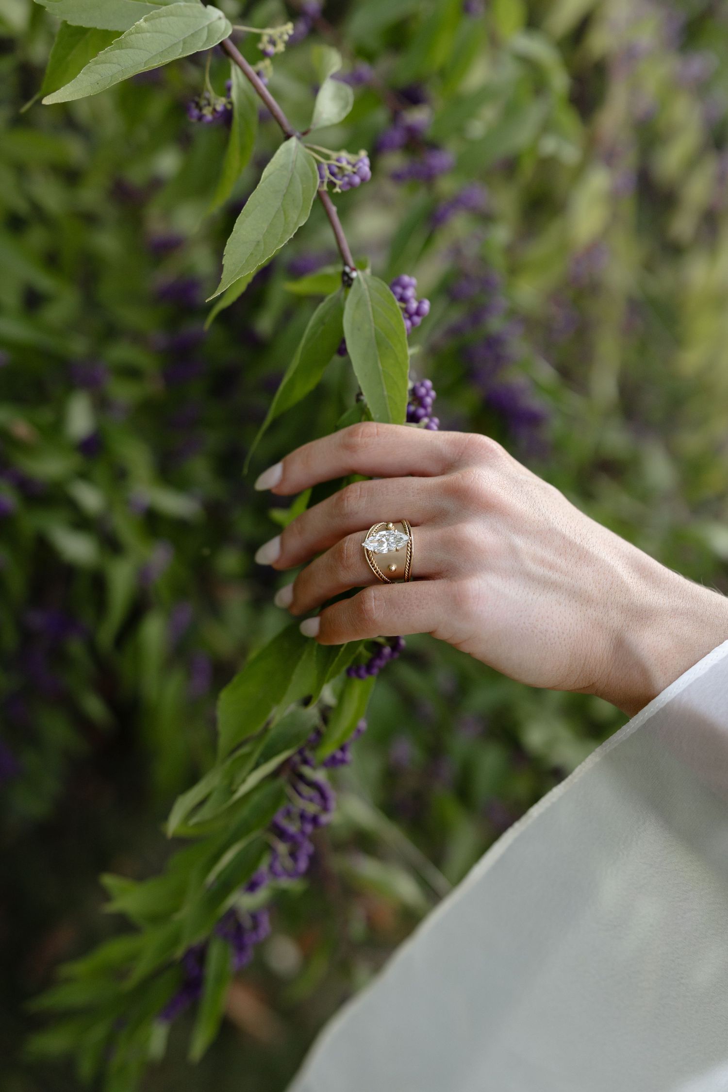 A delicate hand wearing an elegant engagement ring reaches for purple flowers against a soft green garden background.