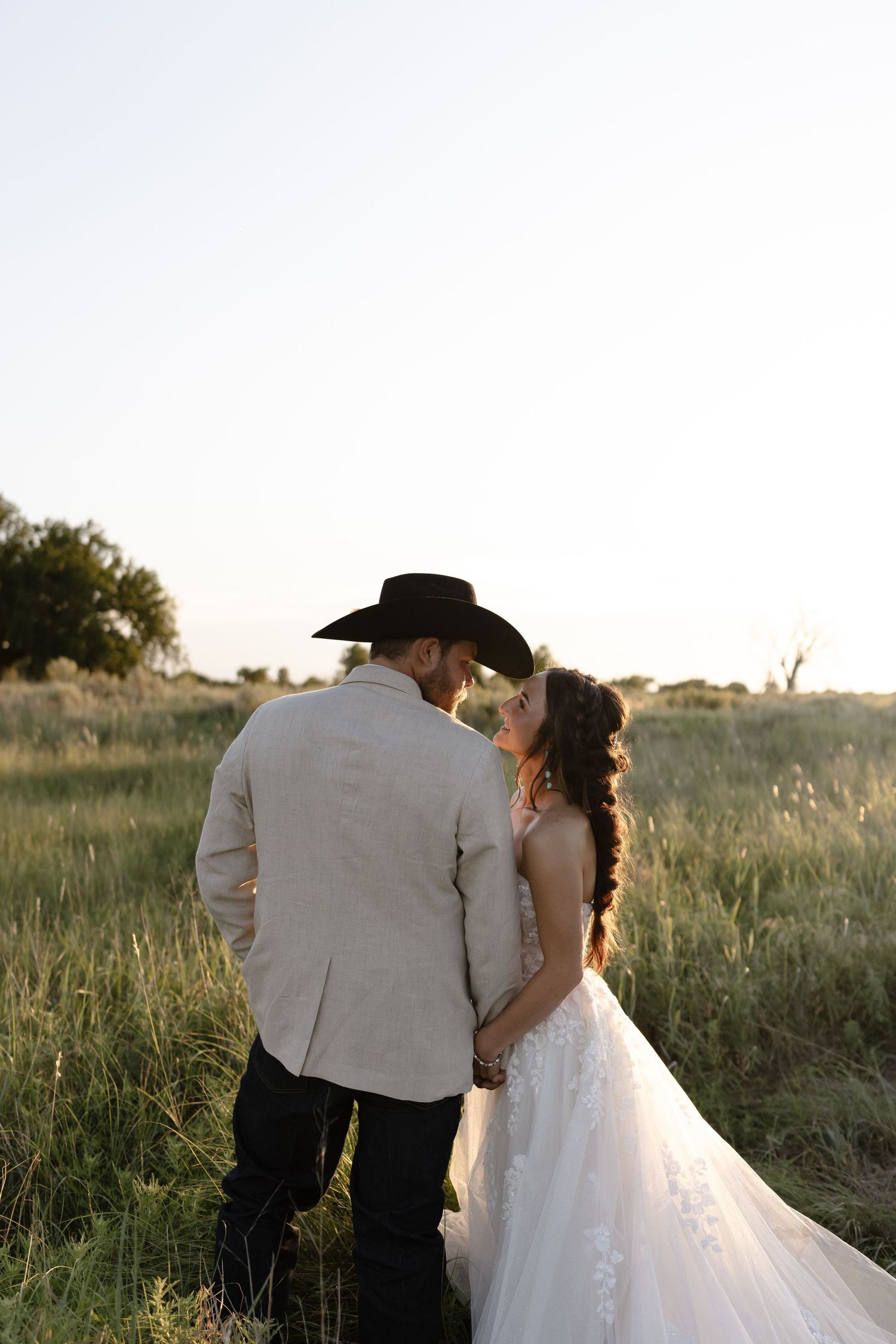 Romantic couple in wedding attire walking through a sunlit meadow at golden hour.