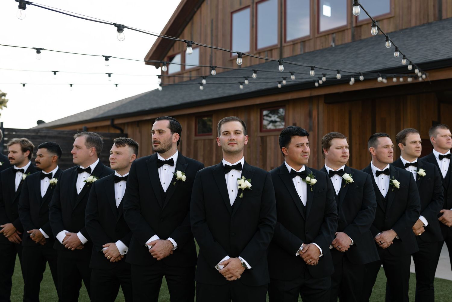 A group of groomsmen in black tuxedos and bow ties stand in a line outside a rustic barn venue with string lights overhead.