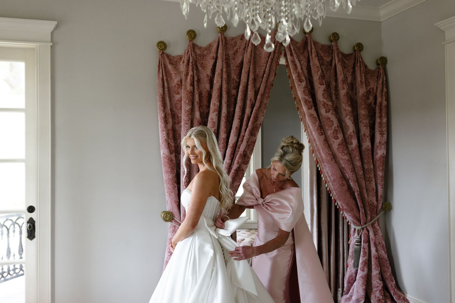 Elegant white living room with crystal chandelier and mauve curtains features ornate crown molding and classic decor.