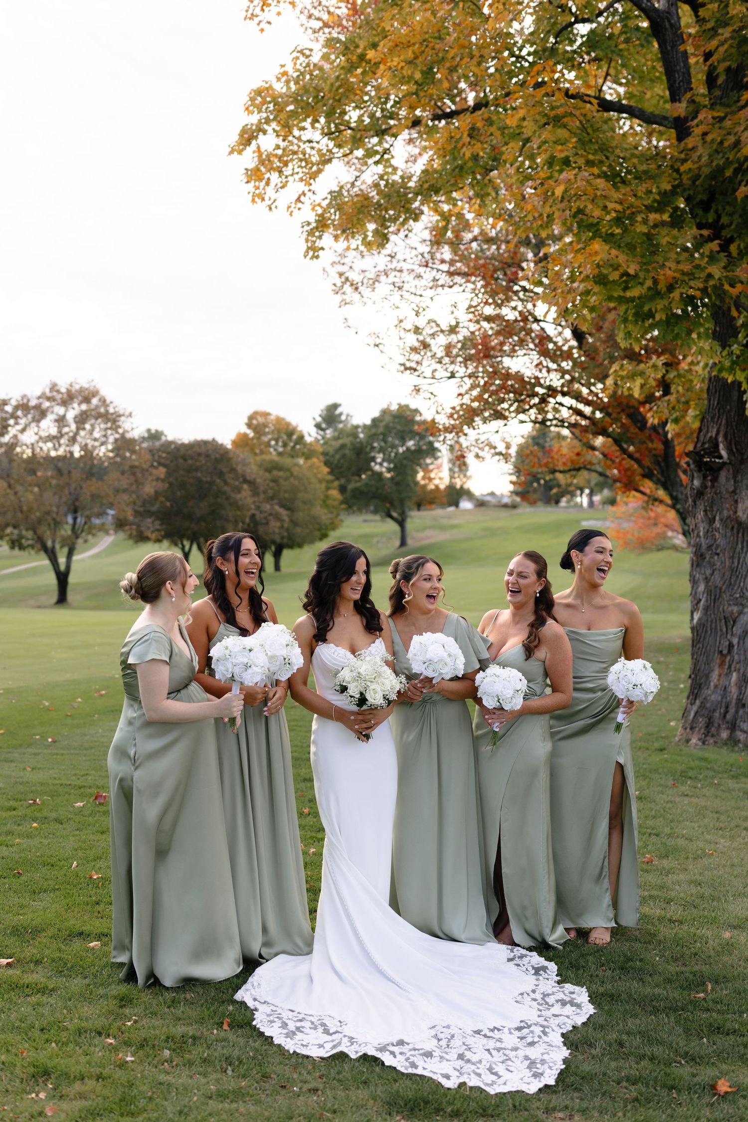 A bridal party in sage green dresses and white gown pose together on a grassy hillside during autumn.