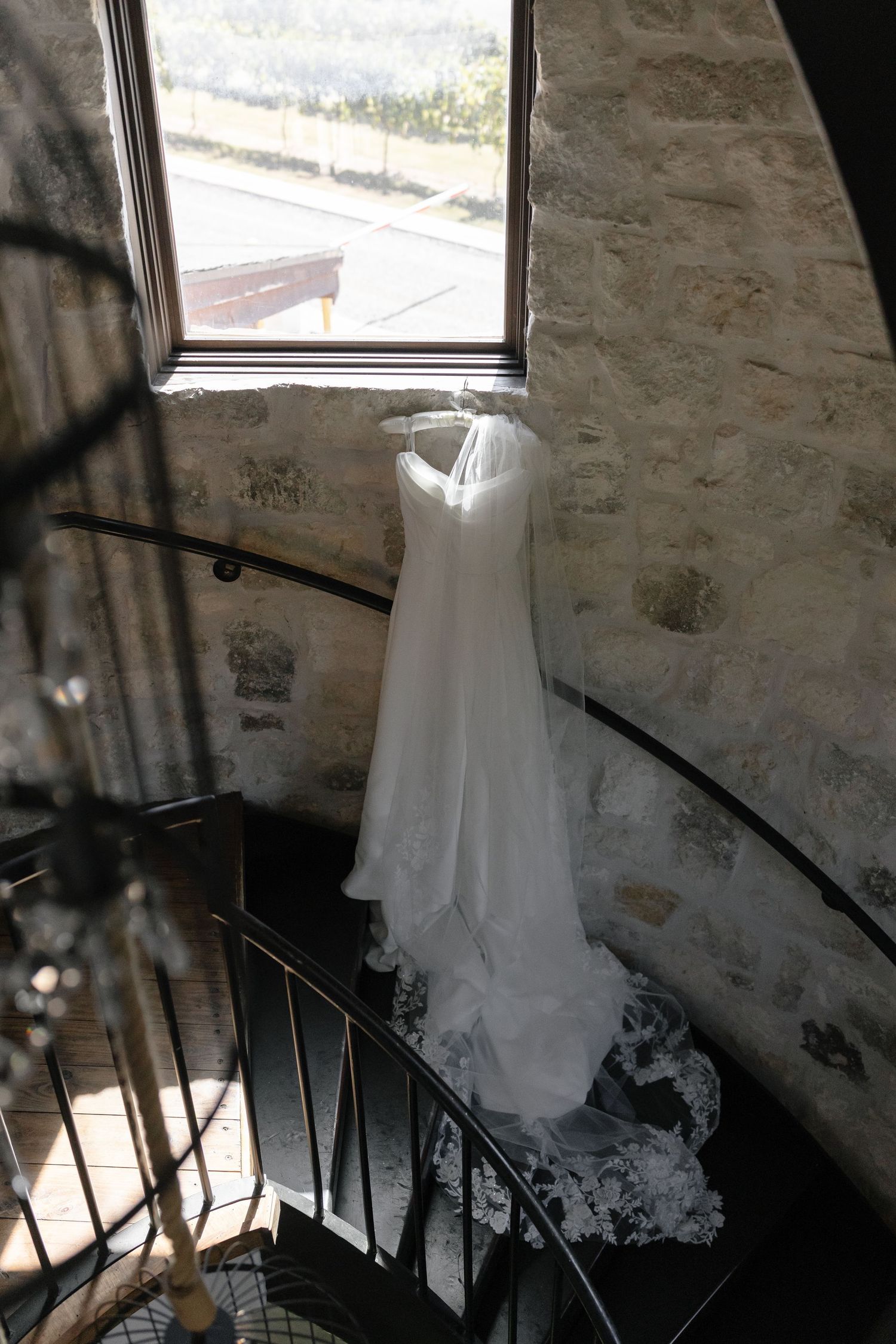 A white wedding dress hangs from a staircase railing in a rustic stone building with natural light streaming through windows.