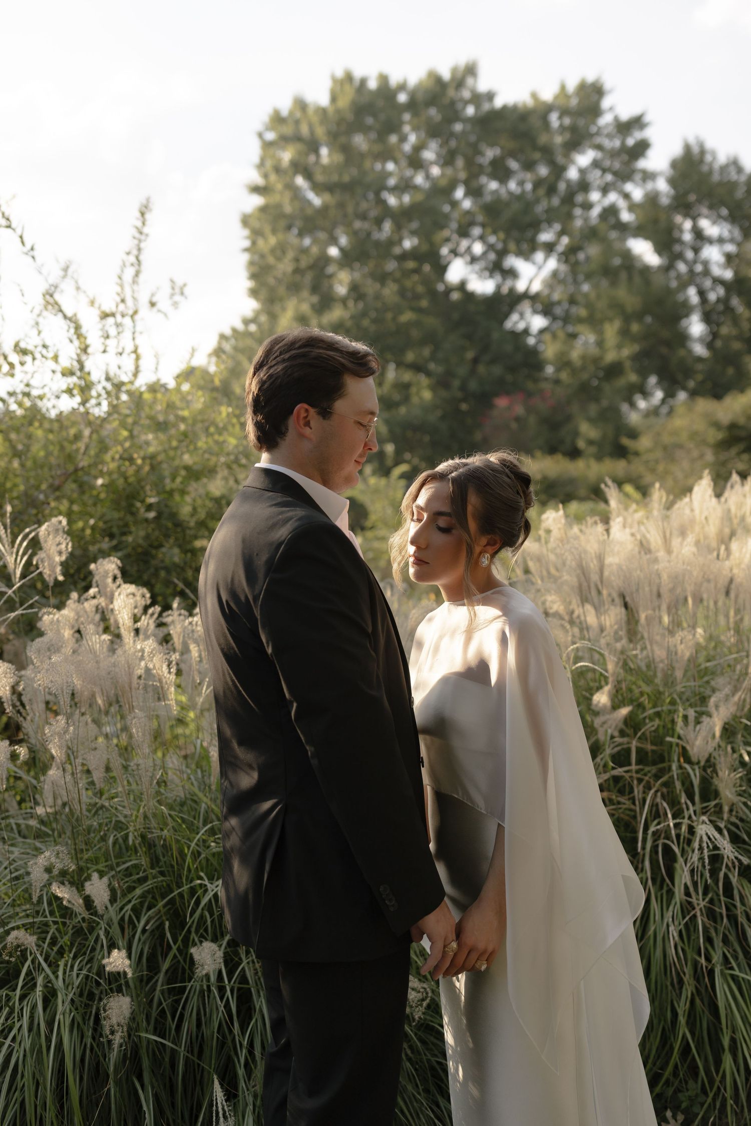 A couple in wedding attire stands together in a field of tall grasses and pampas during golden hour.