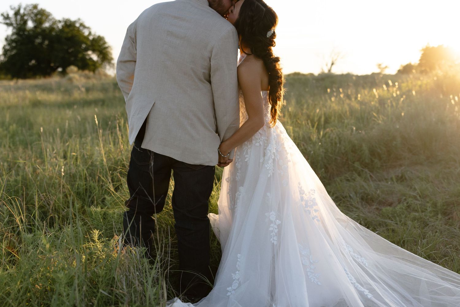 A couple in wedding attire embraces during golden hour in a scenic field at sunset.