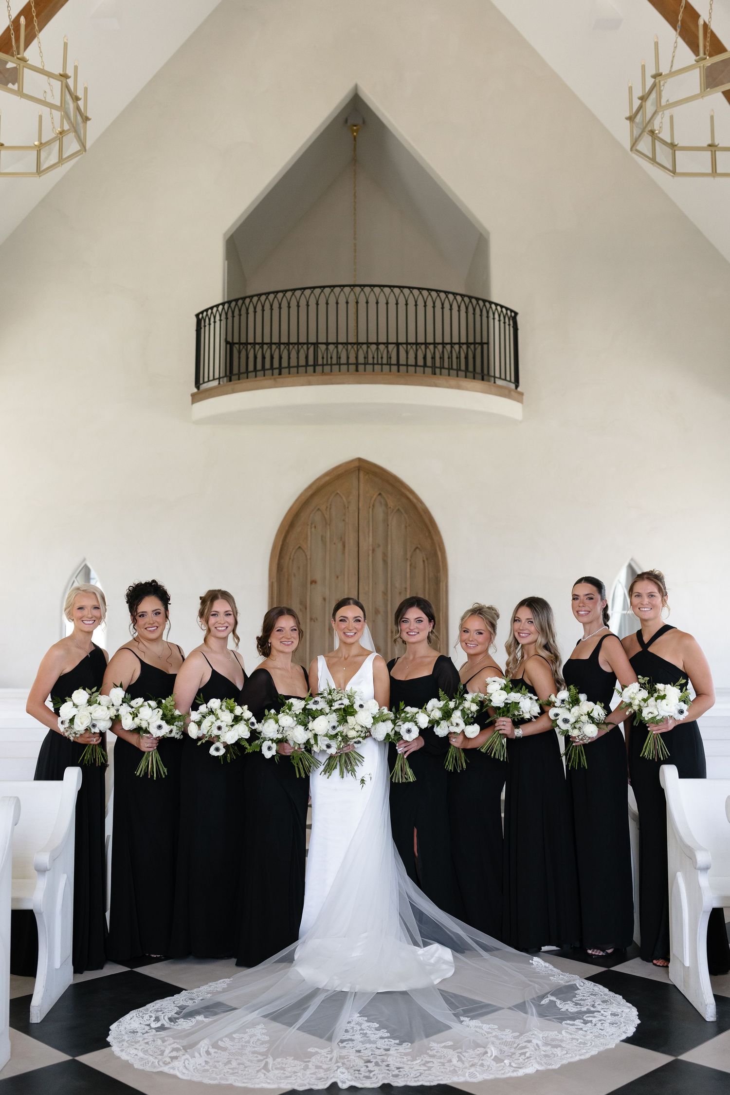 Wedding party in black dresses and white gown pose together in front of elegant white church doorway.