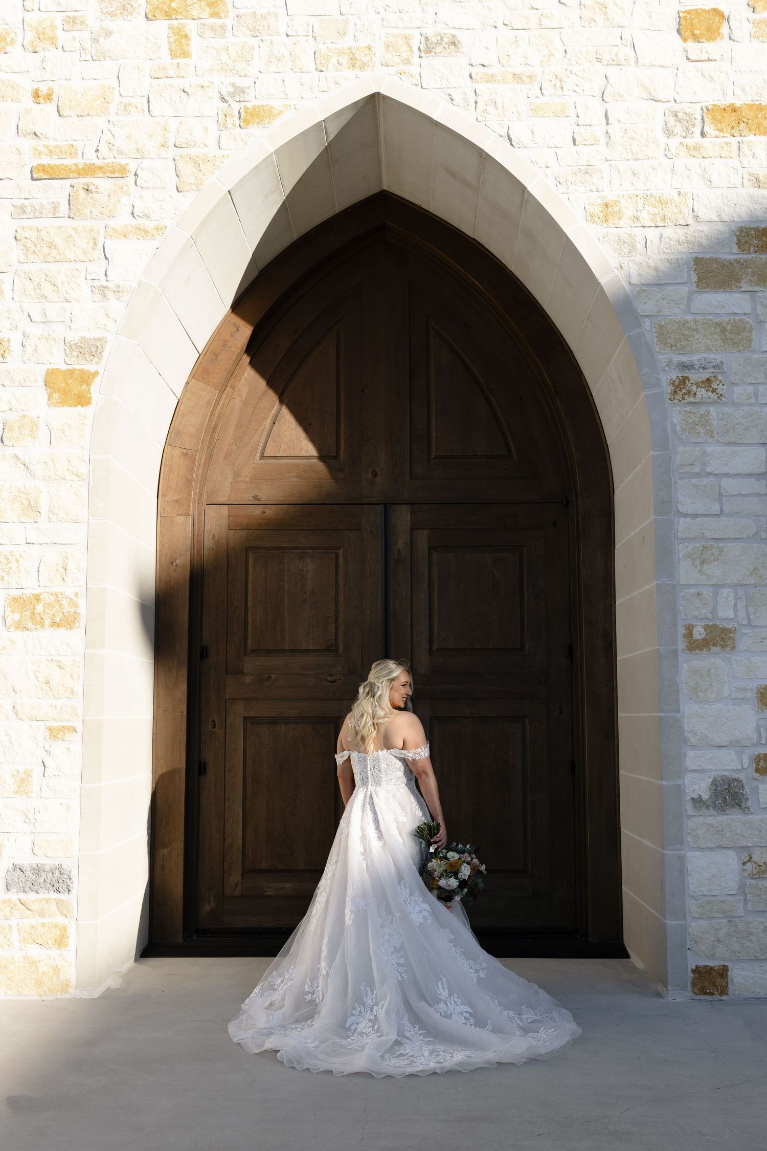 A bride in a flowing white wedding gown poses in front of arched stone doorways at a rustic venue.