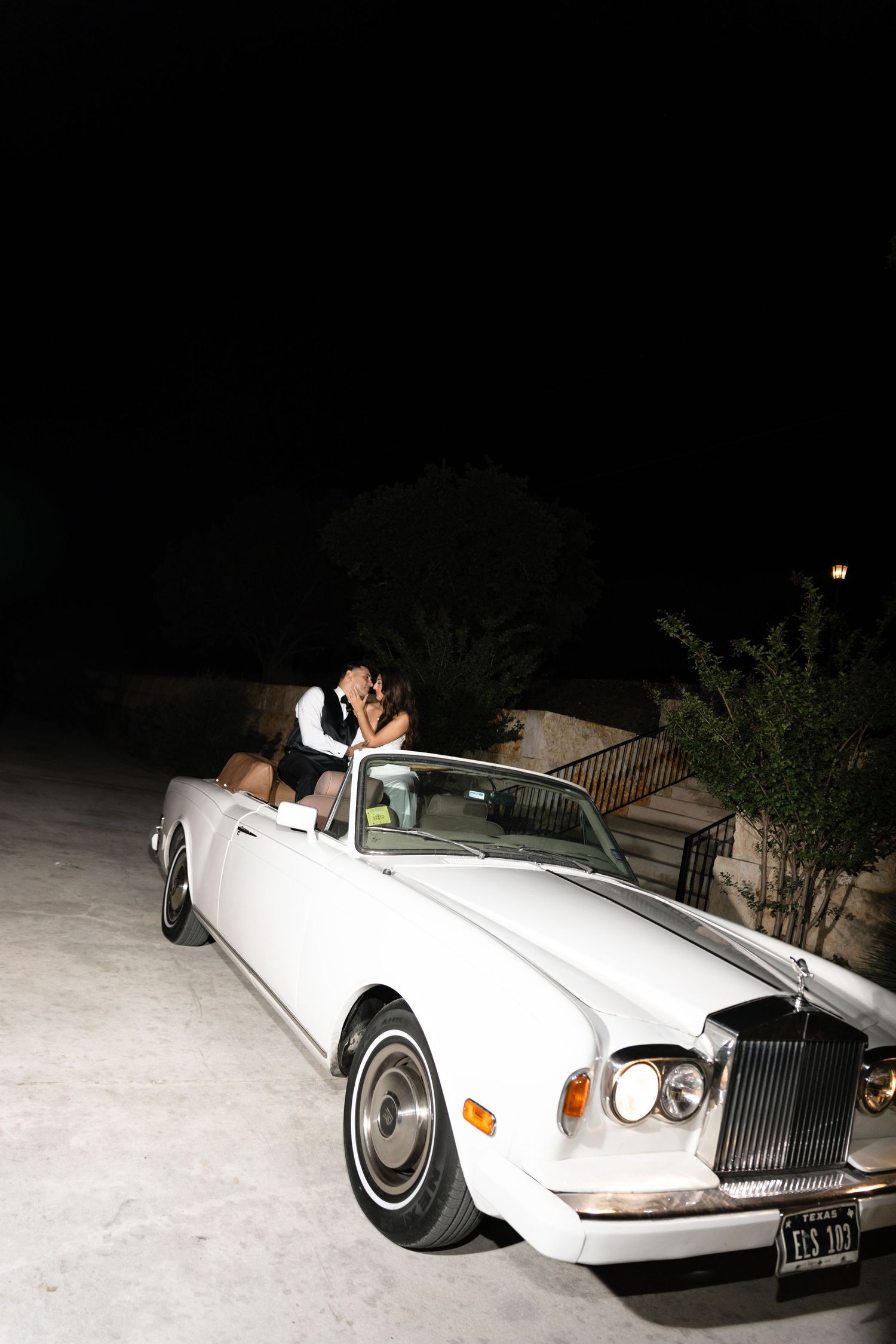 A white vintage Rolls Royce convertible parked at night with its occupants visible in the moonlight.