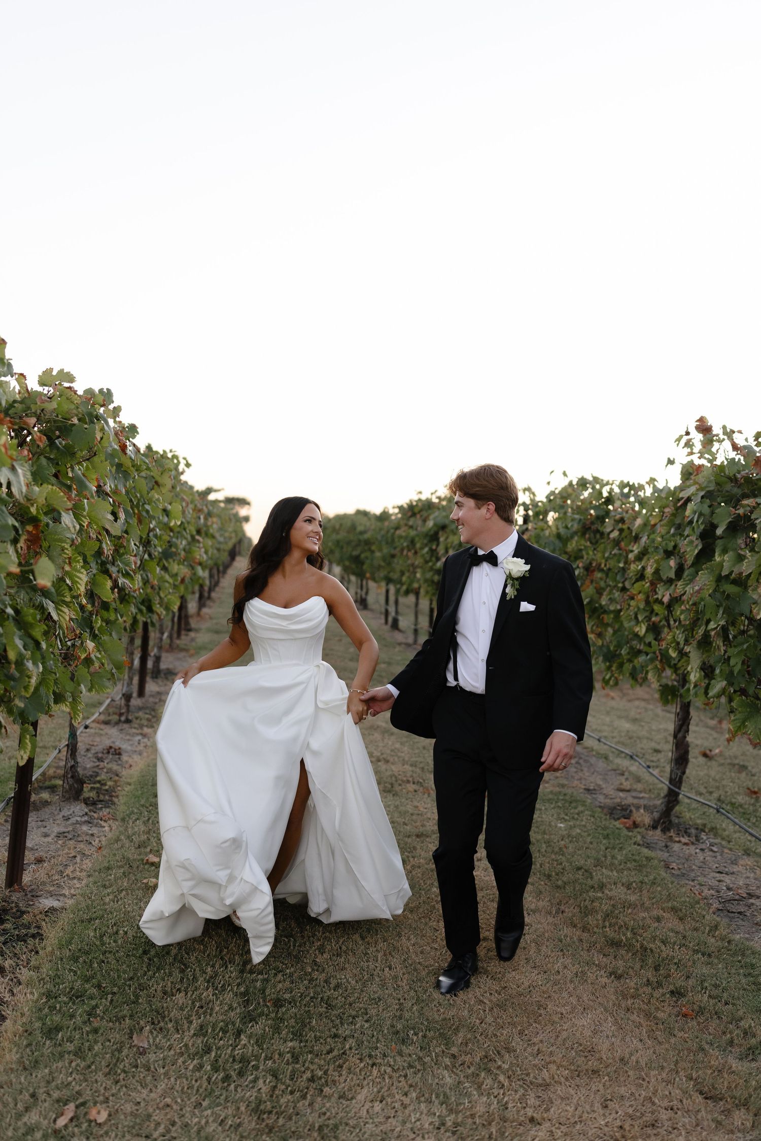 A couple in wedding attire walk together through a scenic vineyard at dusk.