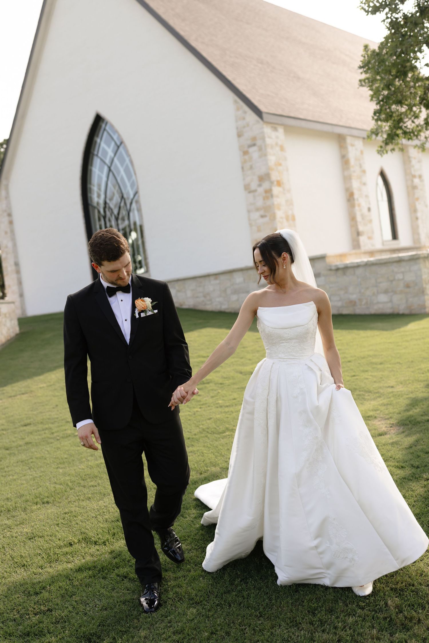 Bride in strapless white gown and groom in black tuxedo hold hands walking outside a white stone chapel.
