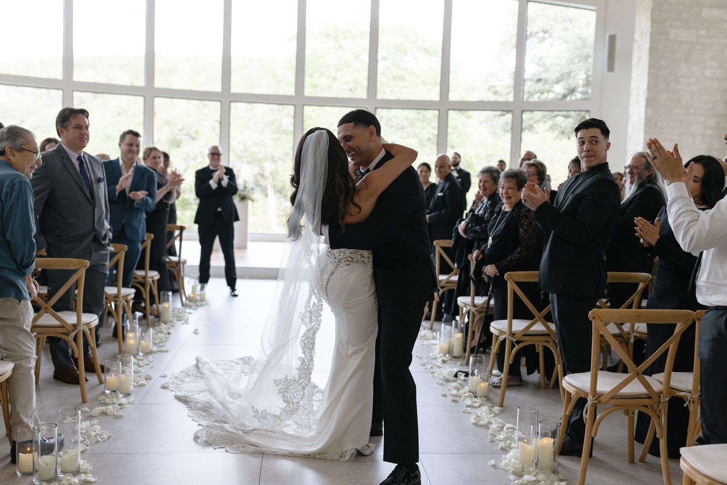 A couple shares their first kiss during an indoor wedding ceremony while guests look on from wooden chairs.