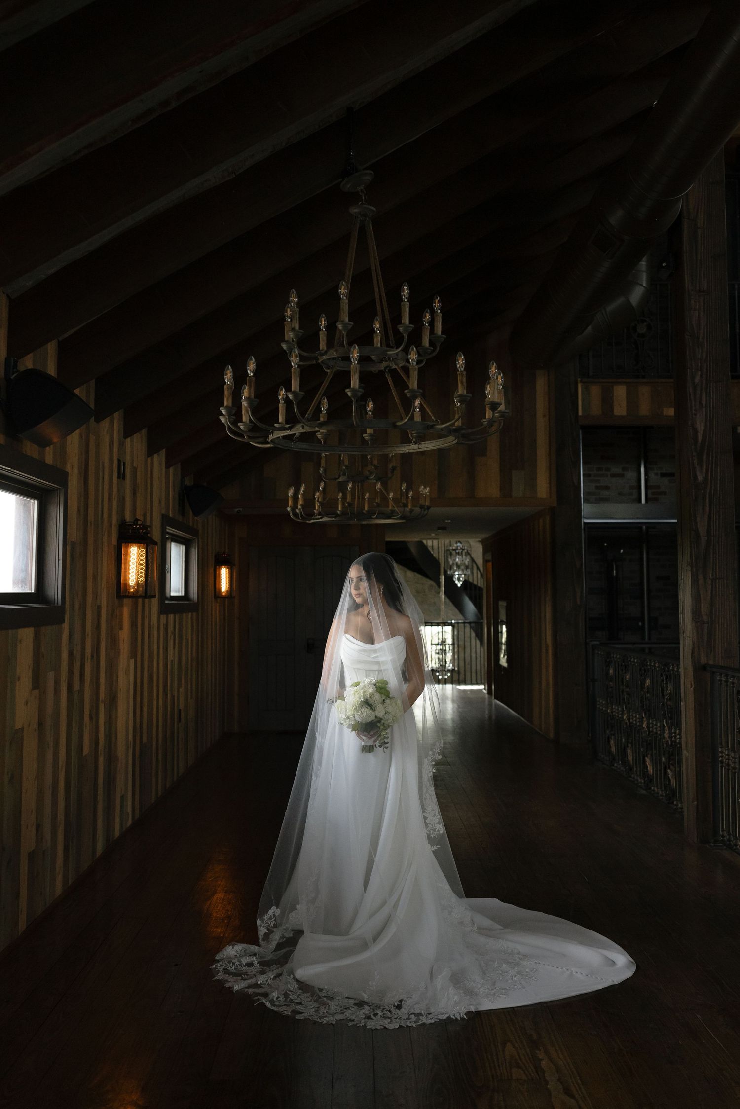 A bride in a flowing white wedding gown and veil stands in a rustic wooden barn venue with dramatic lighting.