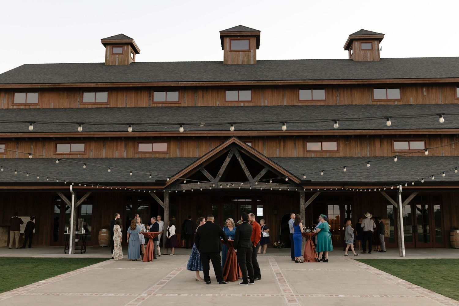 People gather outside a rustic wooden barn-style venue with multiple chimneys during an outdoor event.