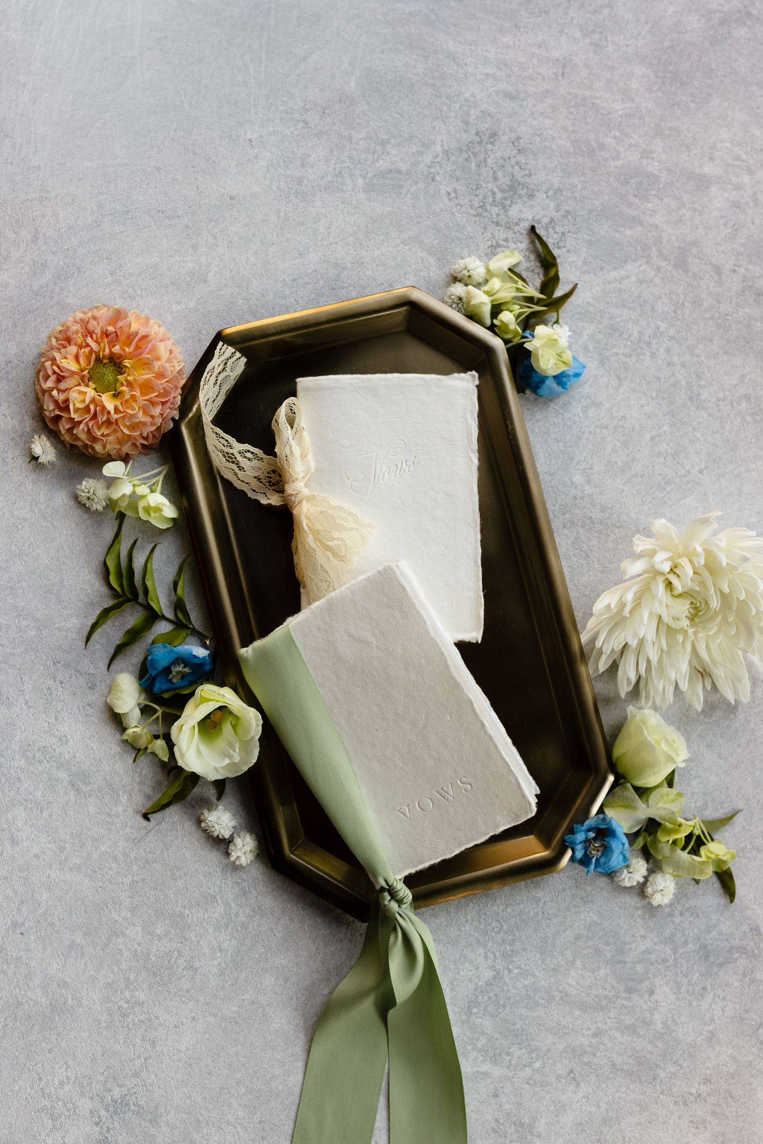 Elegant wedding styling flatlay with white soap bars, delicate flowers, and sage green ribbons on a grey marble surface.