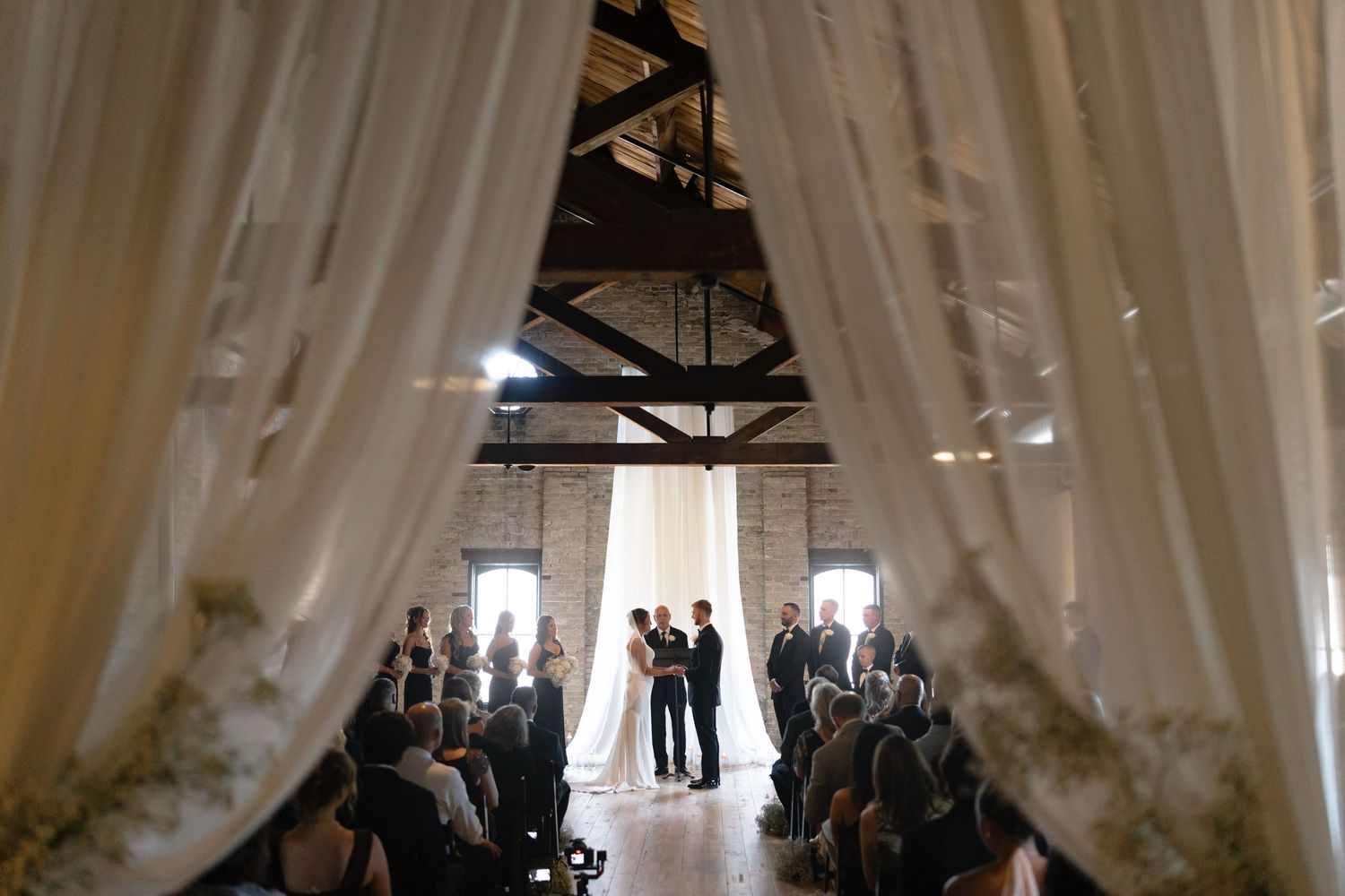 Elegant white fabric drapes create a dramatic ceiling canopy in an industrial-style event space.