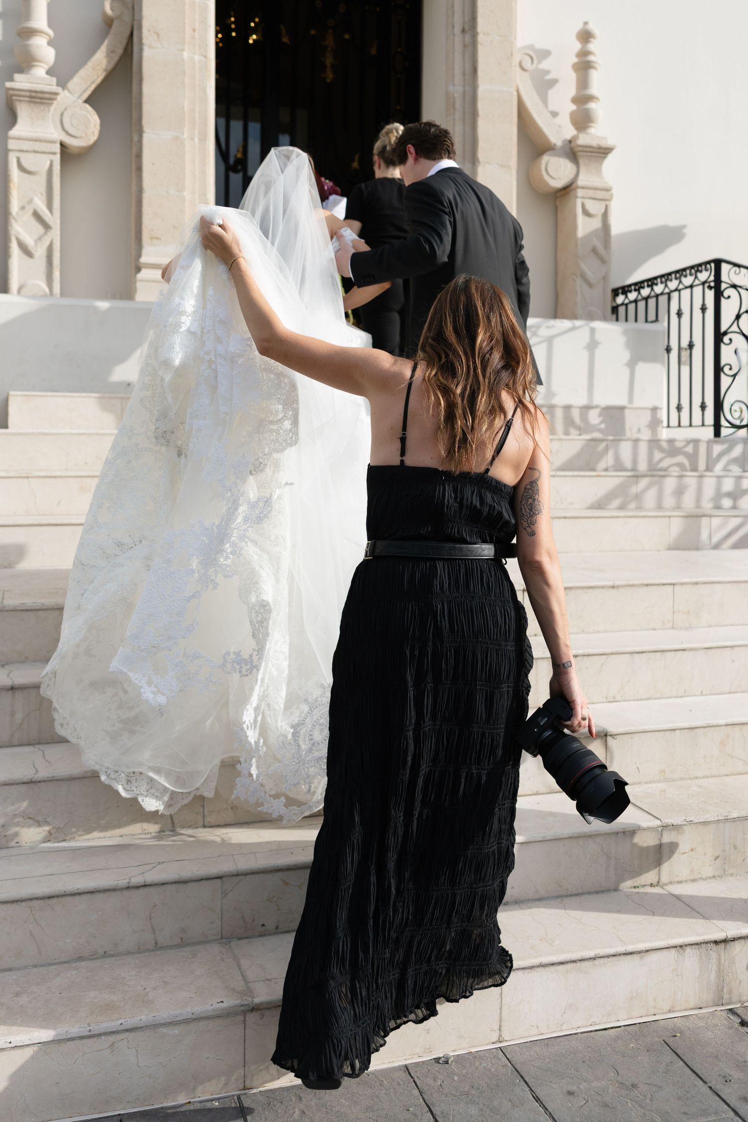 Wedding photographer in black dress carries bride's veil up church steps as couple enters venue.