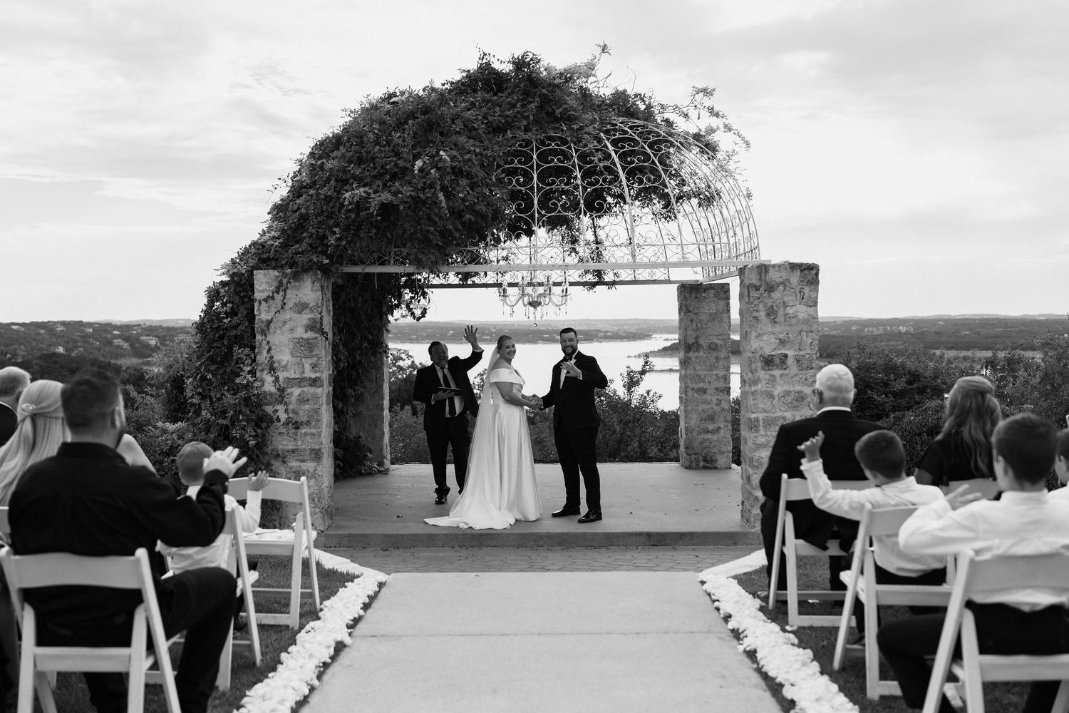 A black and white panoramic view of an outdoor wedding ceremony with guests seated in rows facing a stone structure.