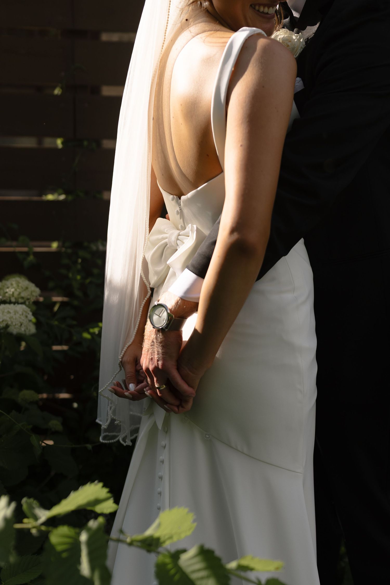 Side view of a bride in a sleek white wedding dress with an open back and wristwatch standing in natural lighting.