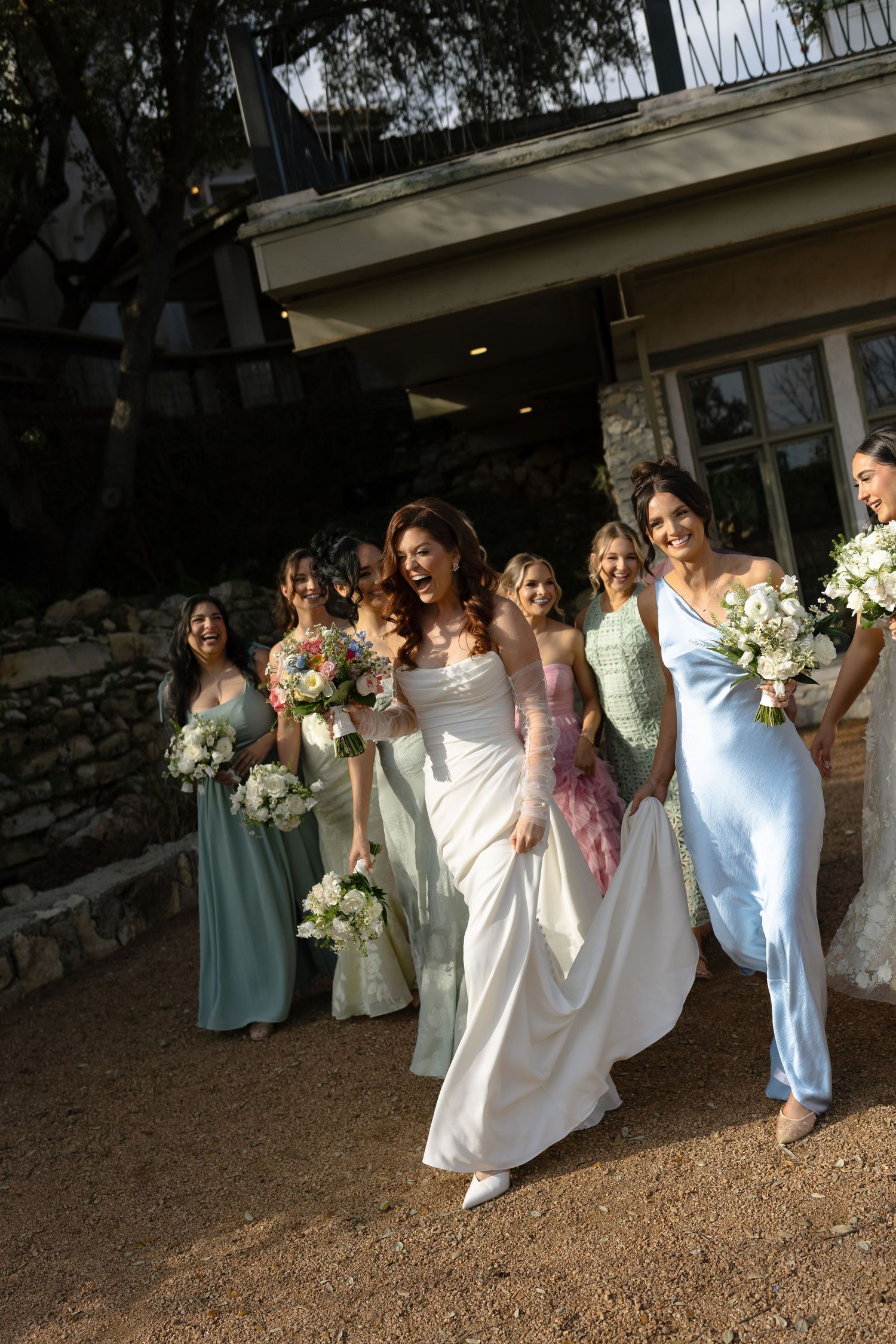 Wedding party in light blue and white dresses pose together on rustic outdoor grounds at sunset.