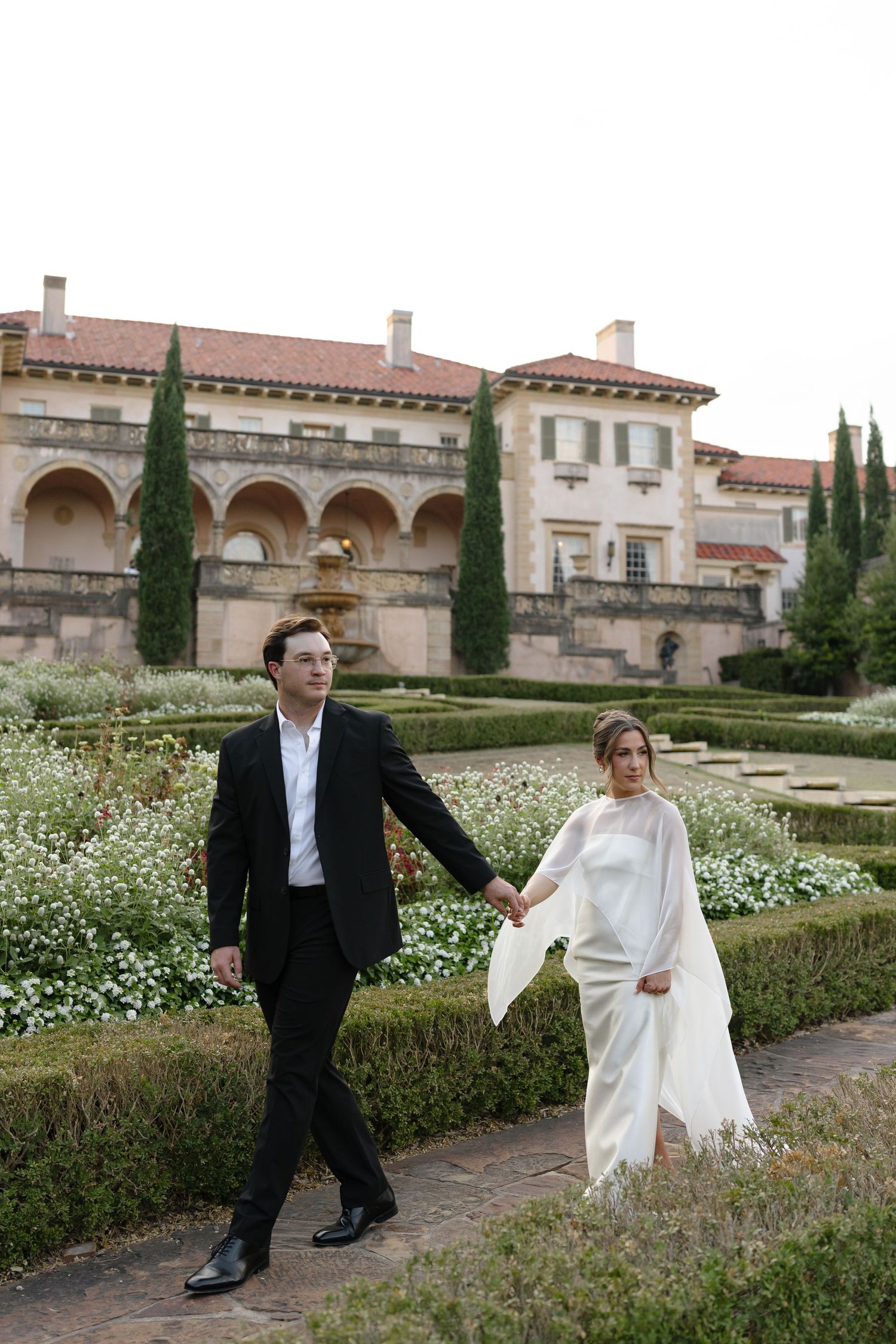 A couple walks through a formal garden with white flowers and cypress trees near an elegant Mediterranean villa.