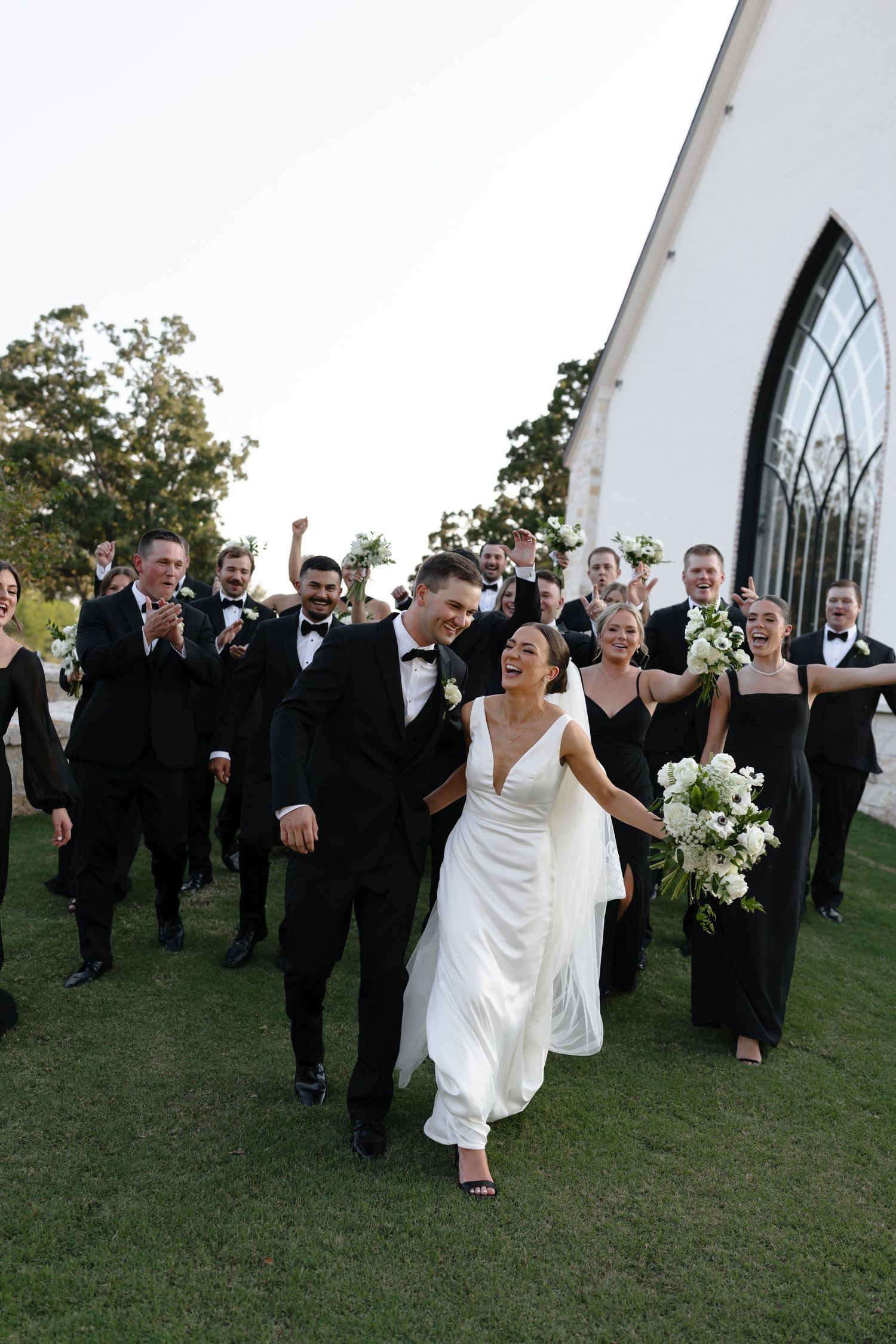 Wedding party in formal attire walking and celebrating on a grassy hill outside.