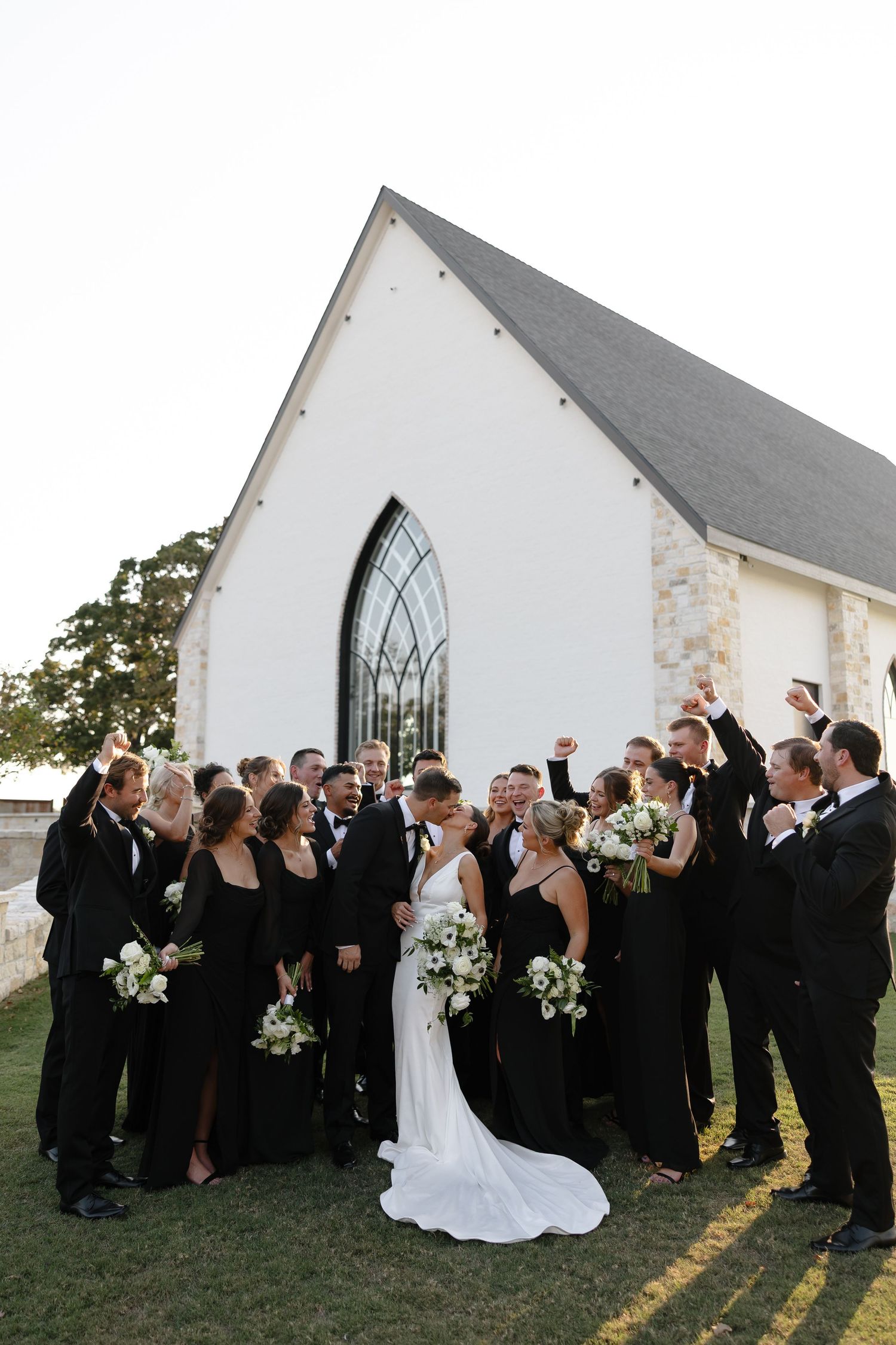 A wedding party in black attire celebrates outside a white church building during a sunny outdoor ceremony.
