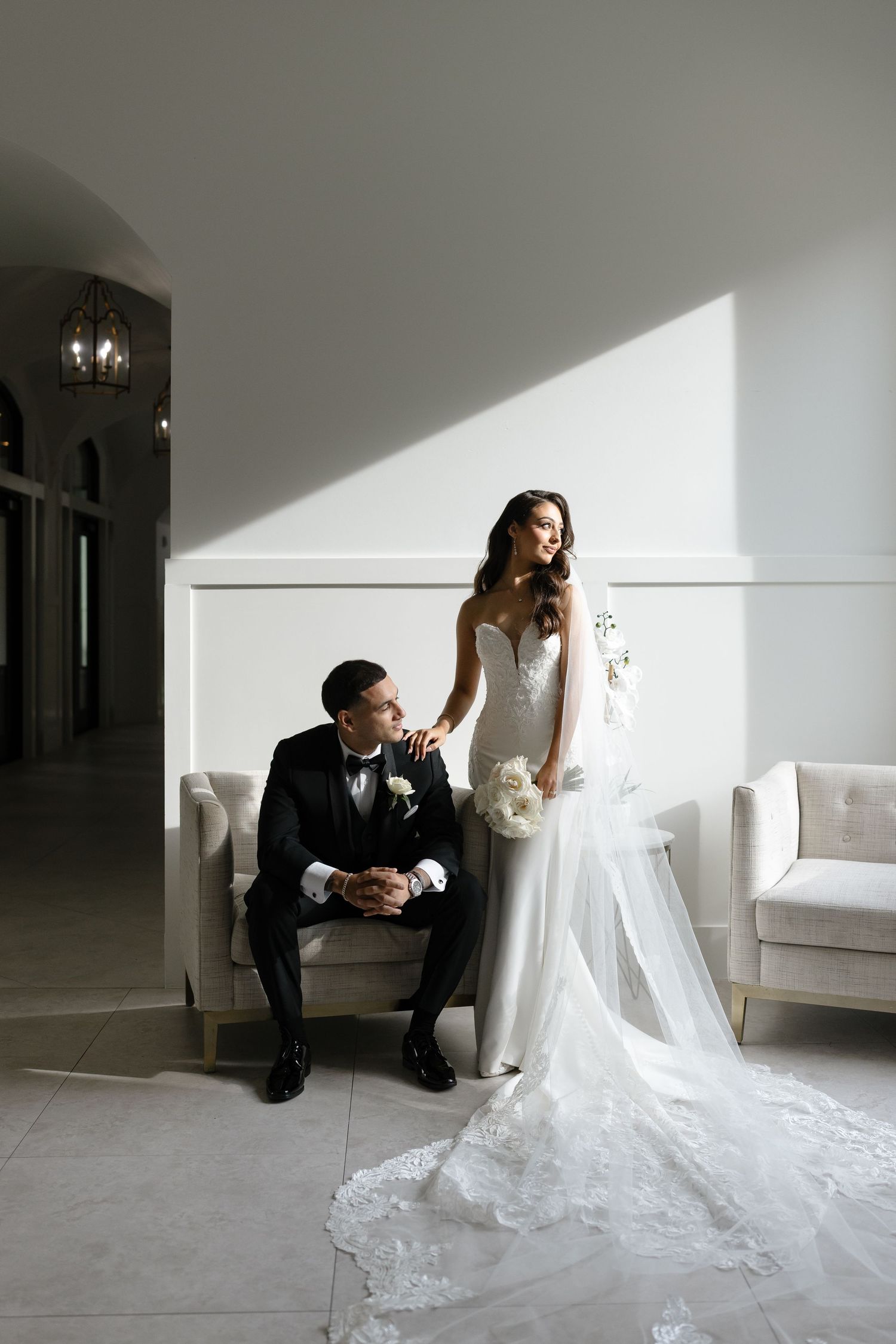 A bride and groom pose together in a minimalist white room with dramatic lighting and shadows.