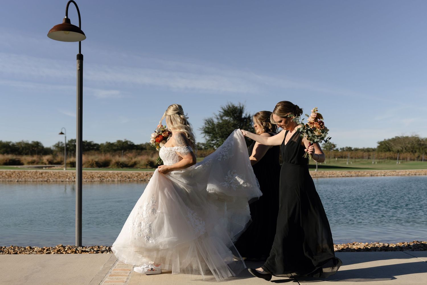 Wedding party in formal attire poses together on a lakeside path with a decorative lamppost and blue sky background.