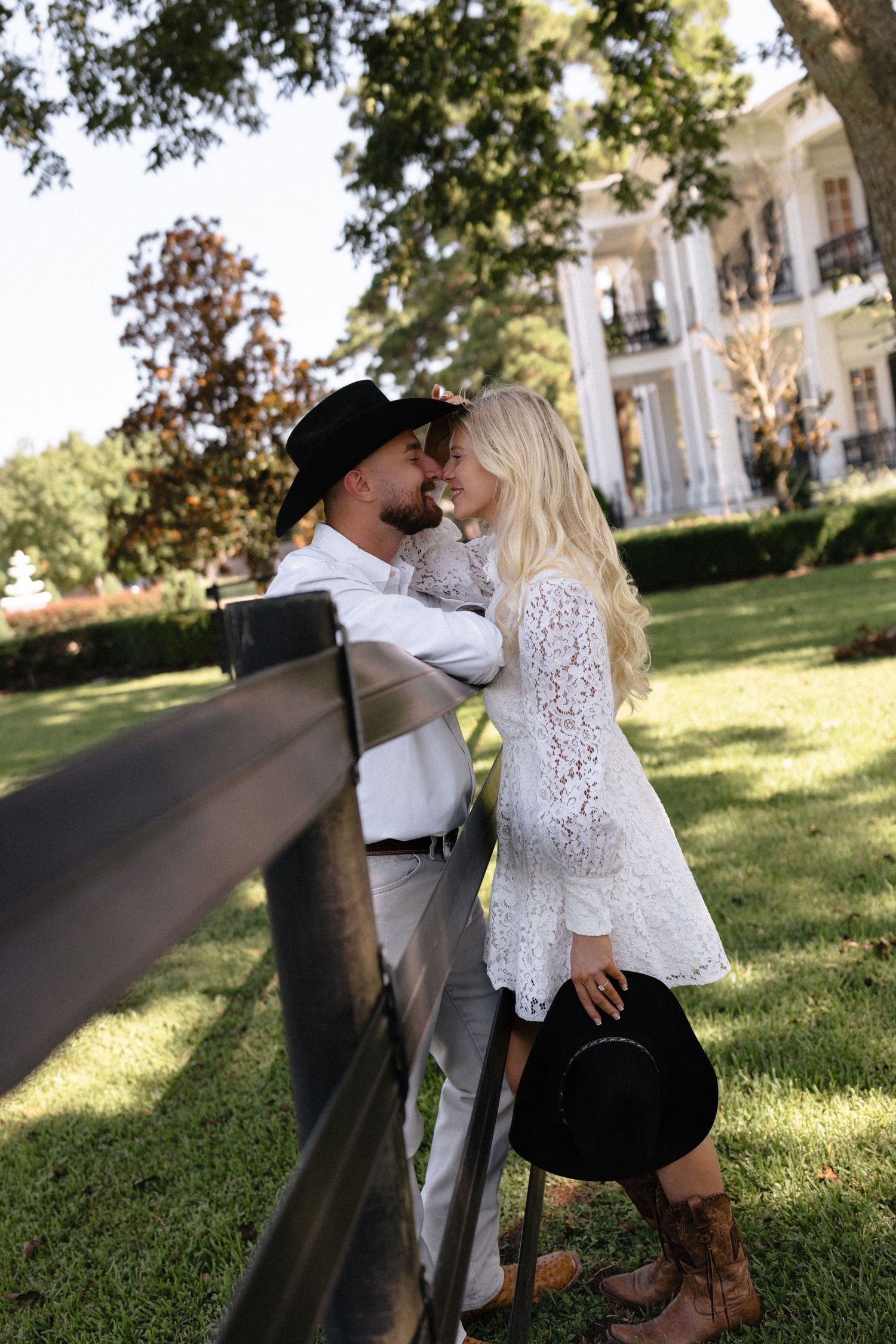 Romantic couple embraces on a sunny lawn with historic buildings in the background during an engagement photoshoot.