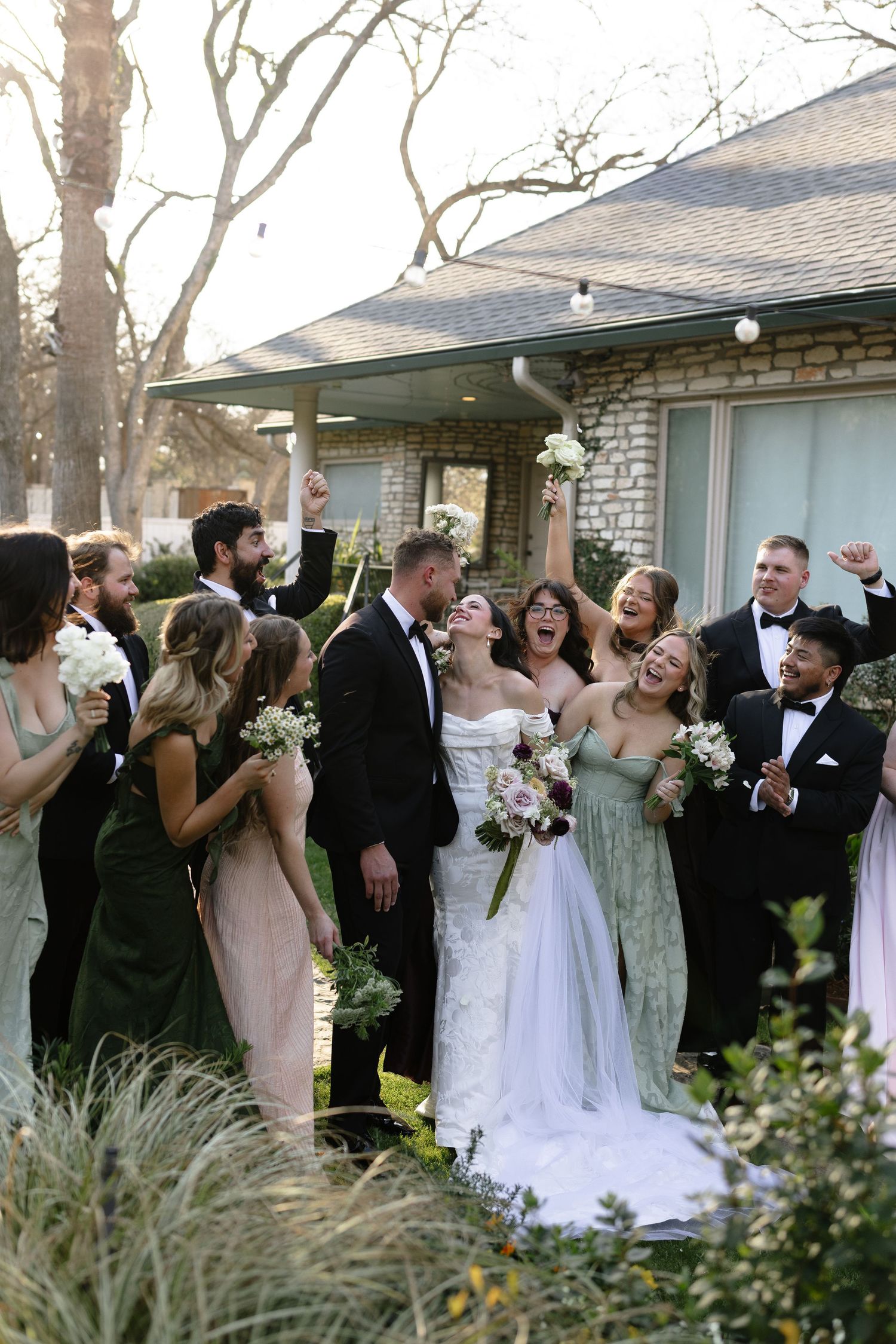 Joyful wedding party celebrating outdoors with bride and groom kissing, bridesmaids in green dresses holding bouquets.