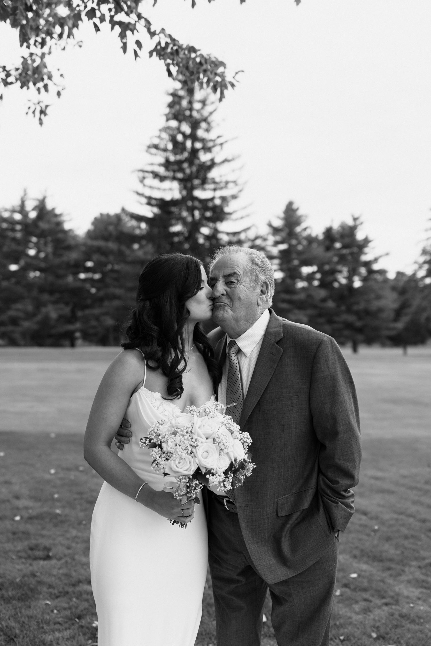 A tender black and white wedding moment captured outdoors with a bouquet of white flowers and evergreen trees in background.