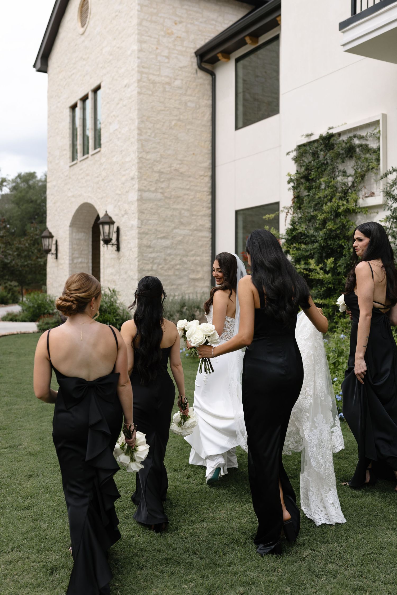 A bride in white and bridesmaids in black dresses stand on a lawn outside an elegant stone and white house.