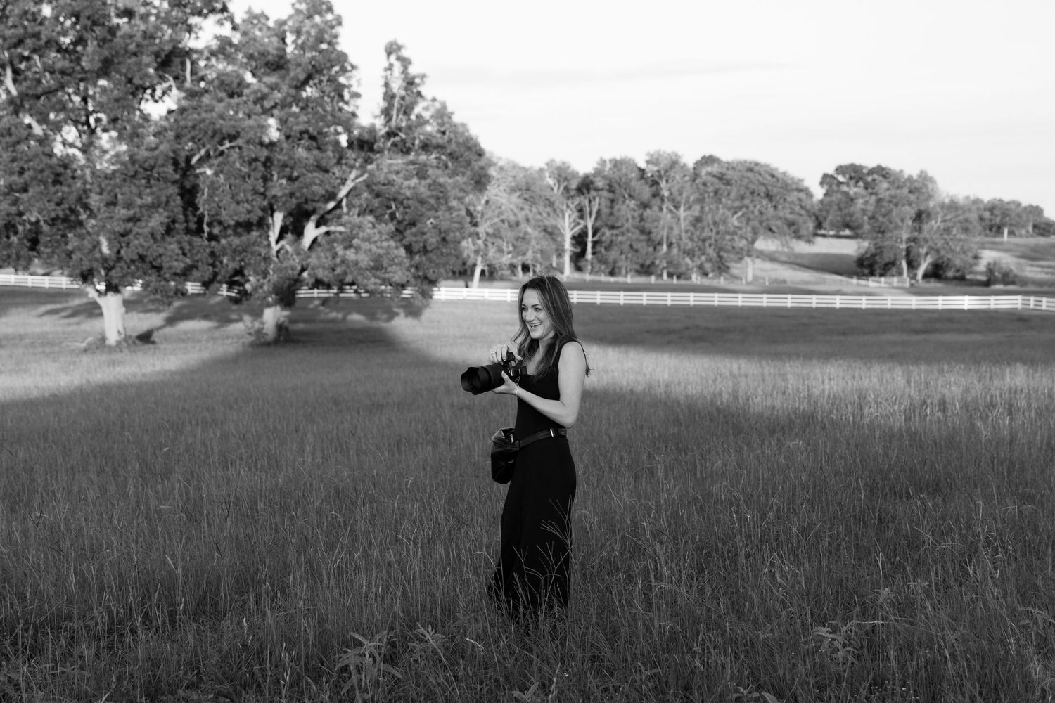Black and white photo of a woman in a field holding a camera, surrounded by tall grass and trees.