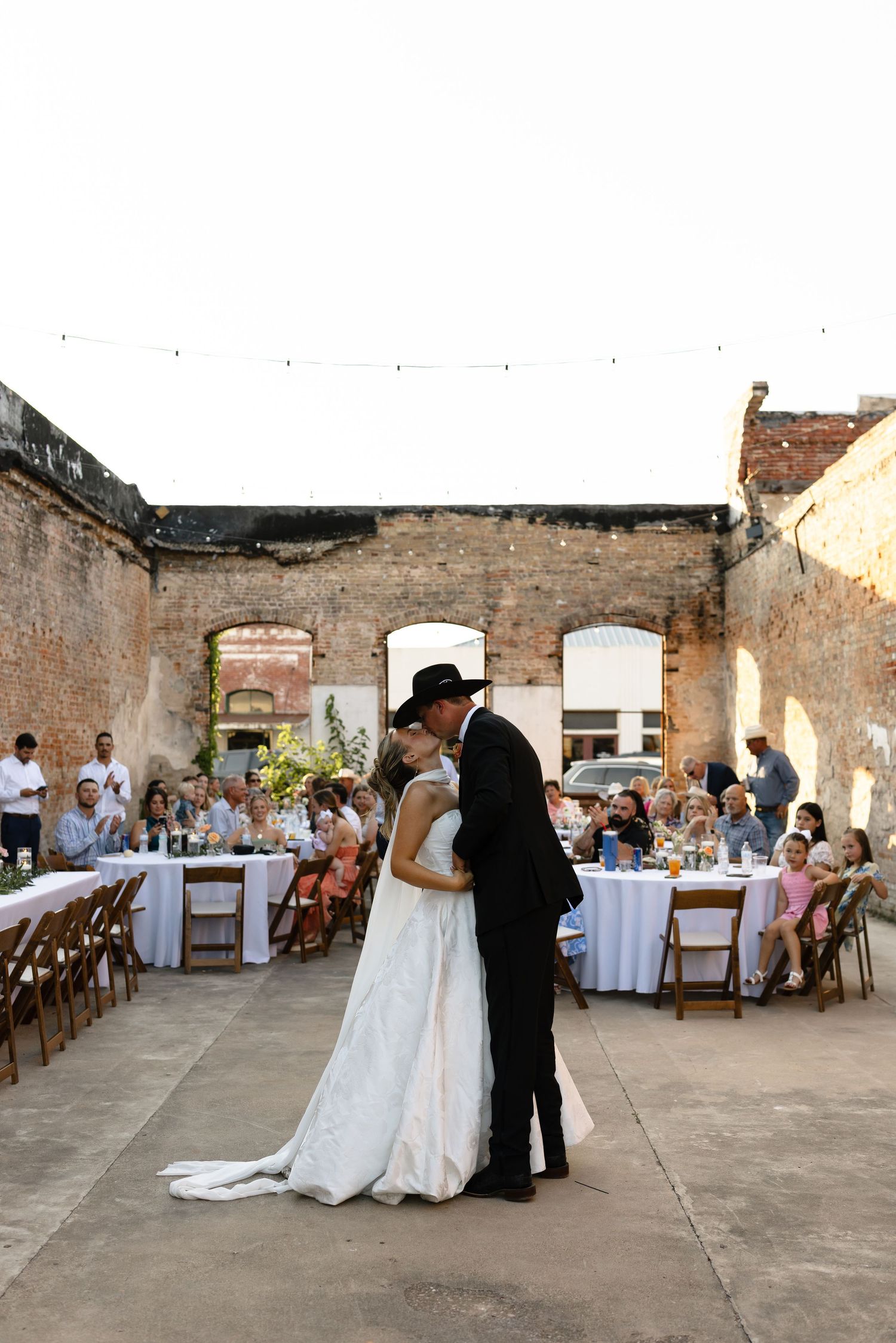 A romantic first dance takes place in a rustic brick-walled venue with elegant table settings and guests looking on.