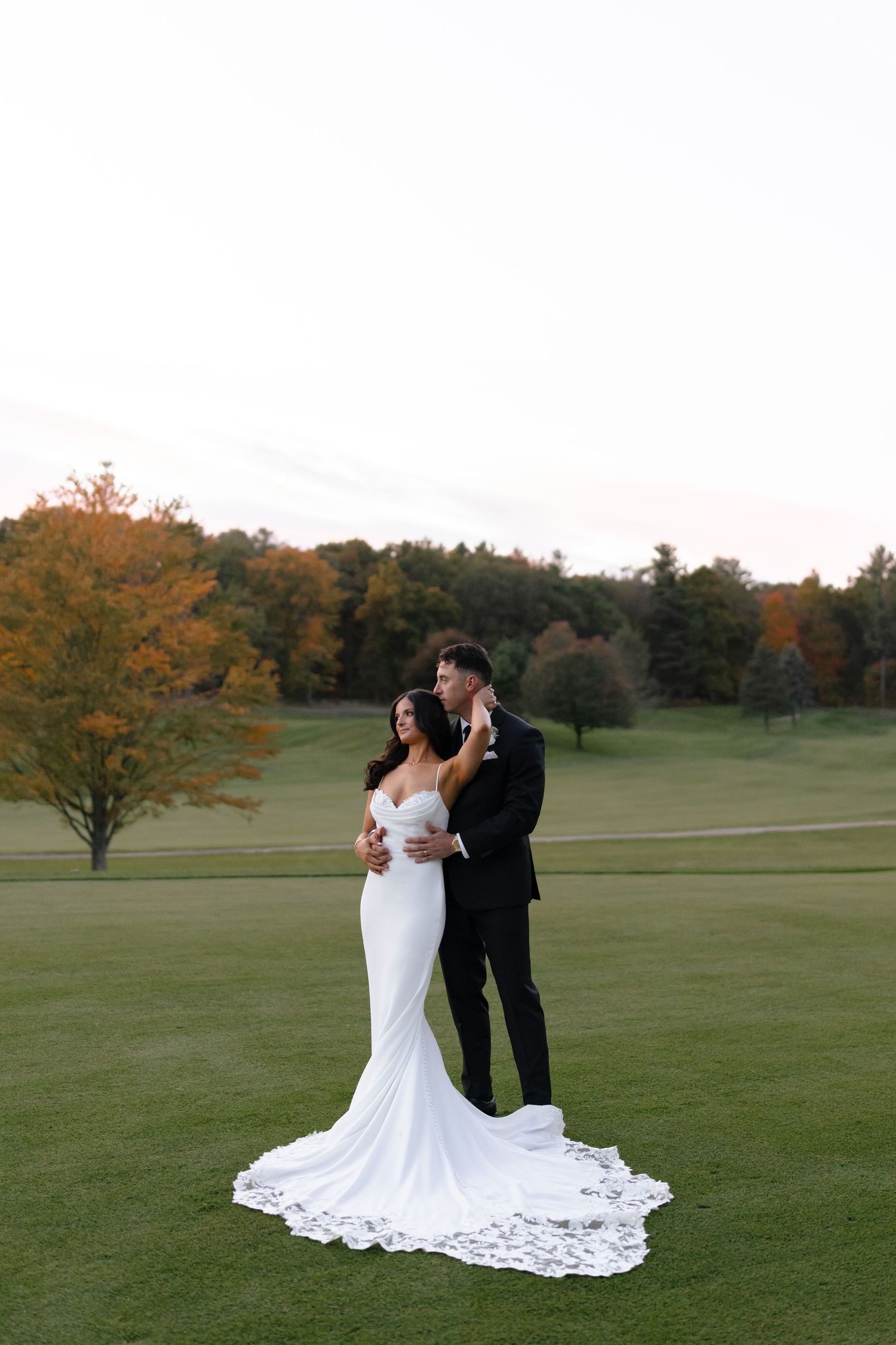 A romantic wedding couple shares an intimate moment on a lush green golf course with autumn trees in the background.