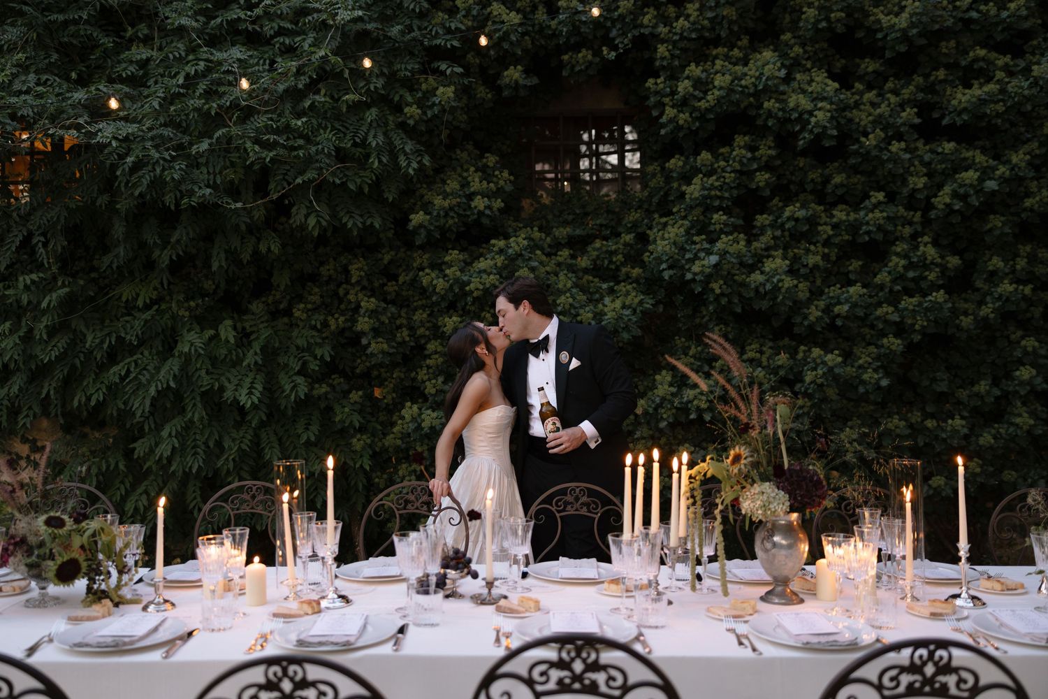 Couple sharing a romantic kiss at an elegantly set outdoor wedding reception table adorned with candles and floral arrangements.