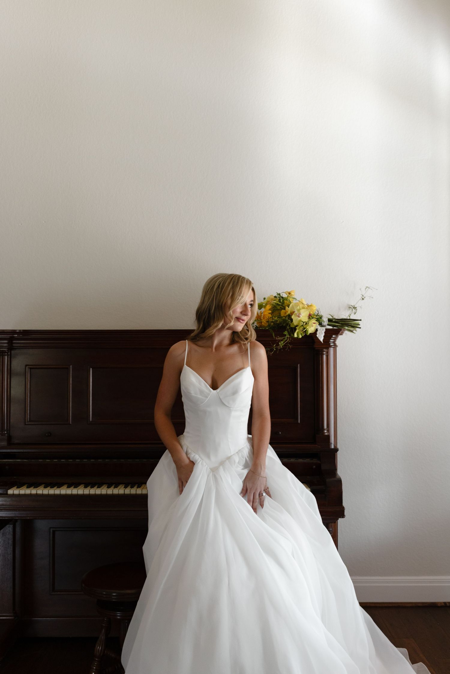 Elegant white wedding gown with spaghetti straps and full tulle skirt shown next to vintage upright piano with floral arrangement.