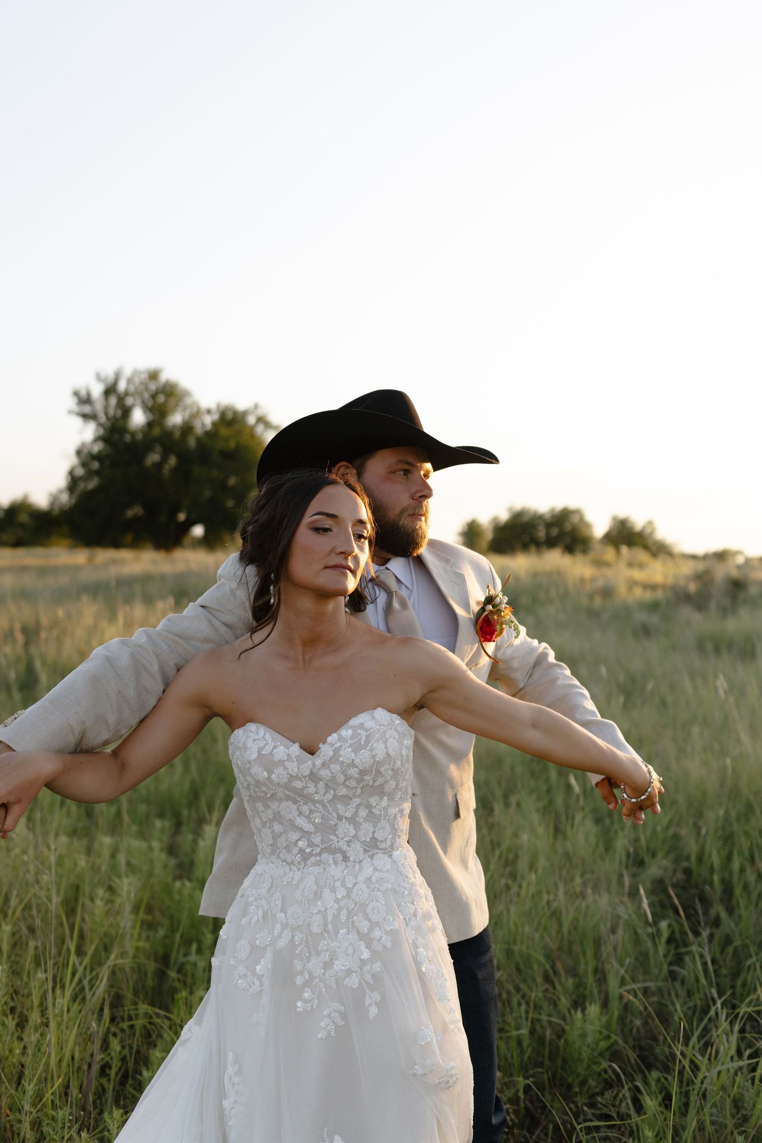 Couple in wedding attire dances playfully together in a golden sunlit field at sunset.