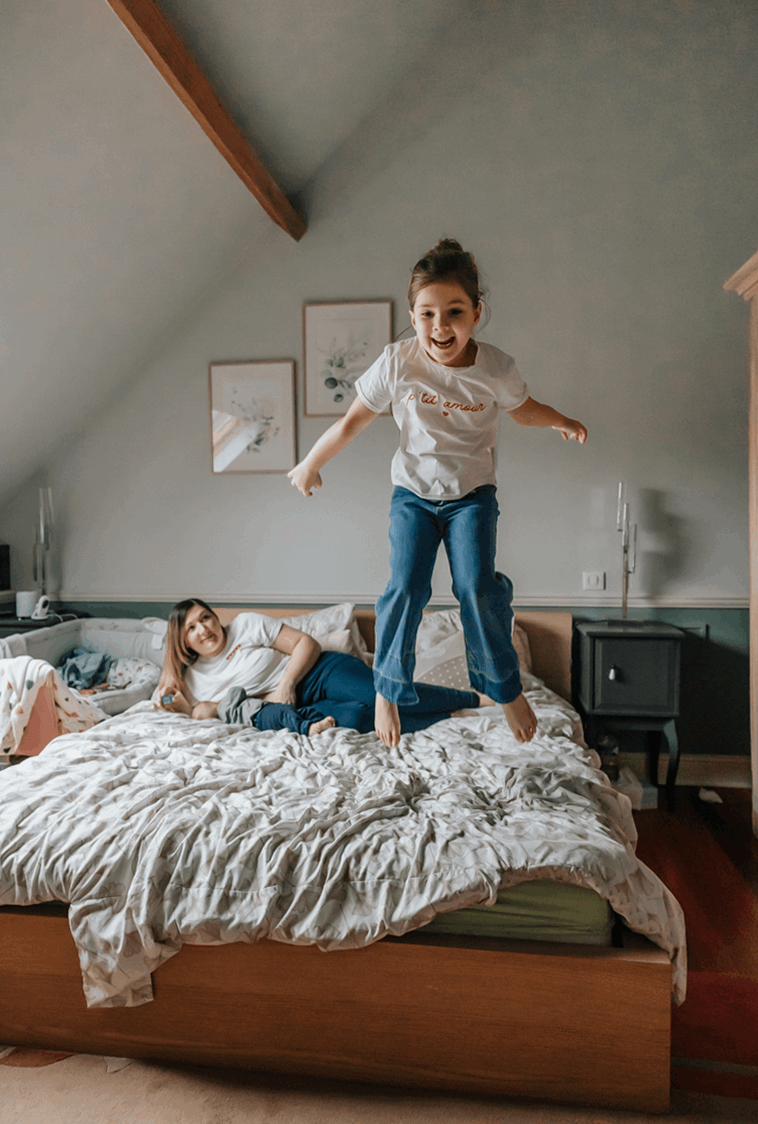 Fille sautant sur le lit pendant que sa maman allaite son petit frere pendant une seance photo de famille a domicile a Lille.