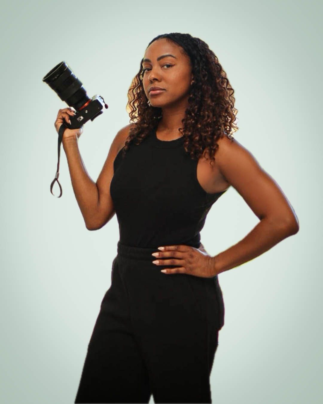 A young Black woman in all-black outfit holds a professional camera, posing confidently against a light background.