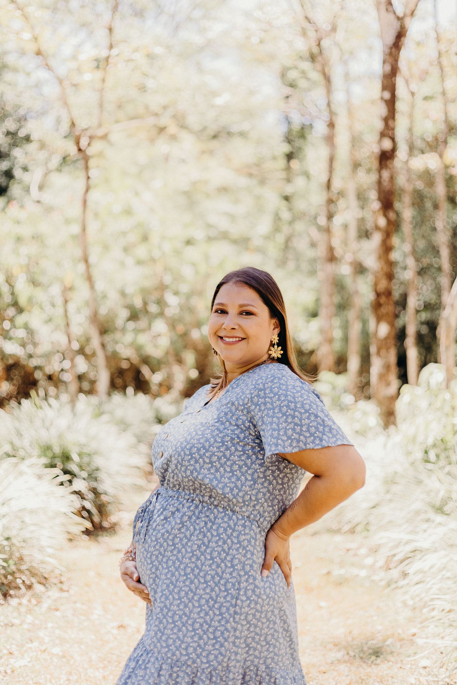 A person in a blue polka dot dress poses outdoors in a sunlit wooded area during a maternity photoshoot.
